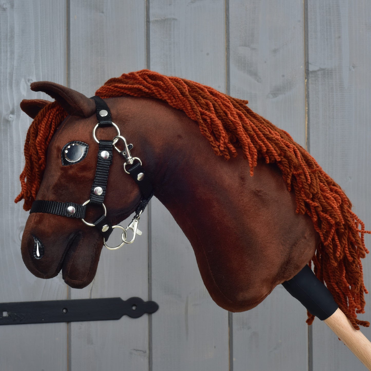 Hobby Horse LarDen "Rebel" velikost XS, s hnědým hannoverským vzhledem, přízovou hřívou, černou ohlávkou a lesklýma očima, držený před šedým dřevěným plotem.
