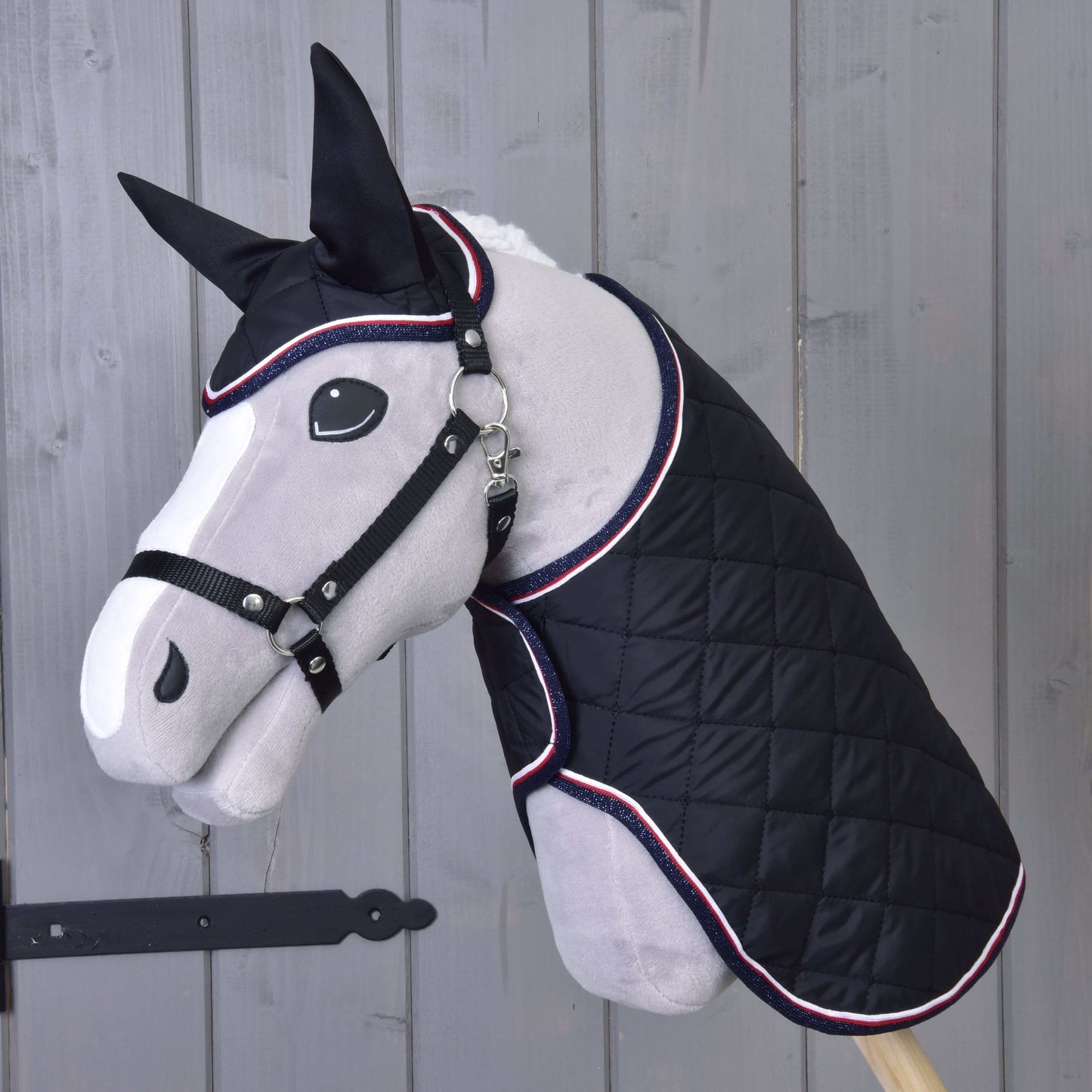 Šedá plyšová figurína koně předvádí Set Royal od Hobby Horse LarDen, černou prošívanou deku a ladící čabraku. V pozadí je světlá dřevěná stěna s černou kovovou zástrčkou vlevo.