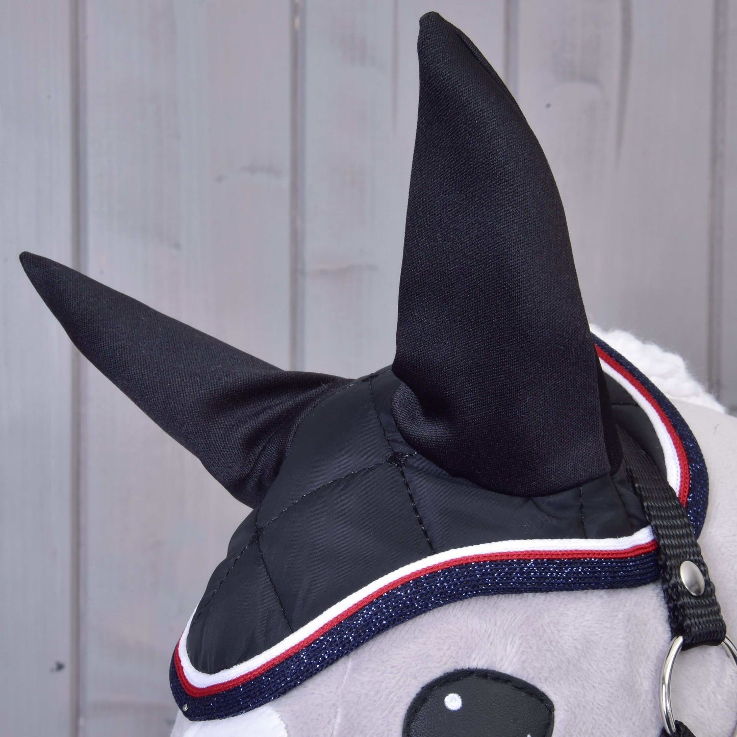 Detail Set Royal od Hobby Horse LarDen: hlava Hobby Horse s černýma látkovýma ušima, stylová čabraka s červeným, bílým a modrým lemem, šedý obličej a černá uzdečka na pozadí světlého dřeva.