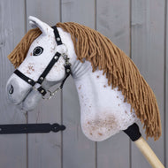 Detail Hobby Horse Rufus s černou ohlávkou od Hobby Horse LarDen s bílým obličejem, hnědou hřívou z příze, černou ohlávkou a hnědými skvrnami, na pozadí dřevěných panelů.
