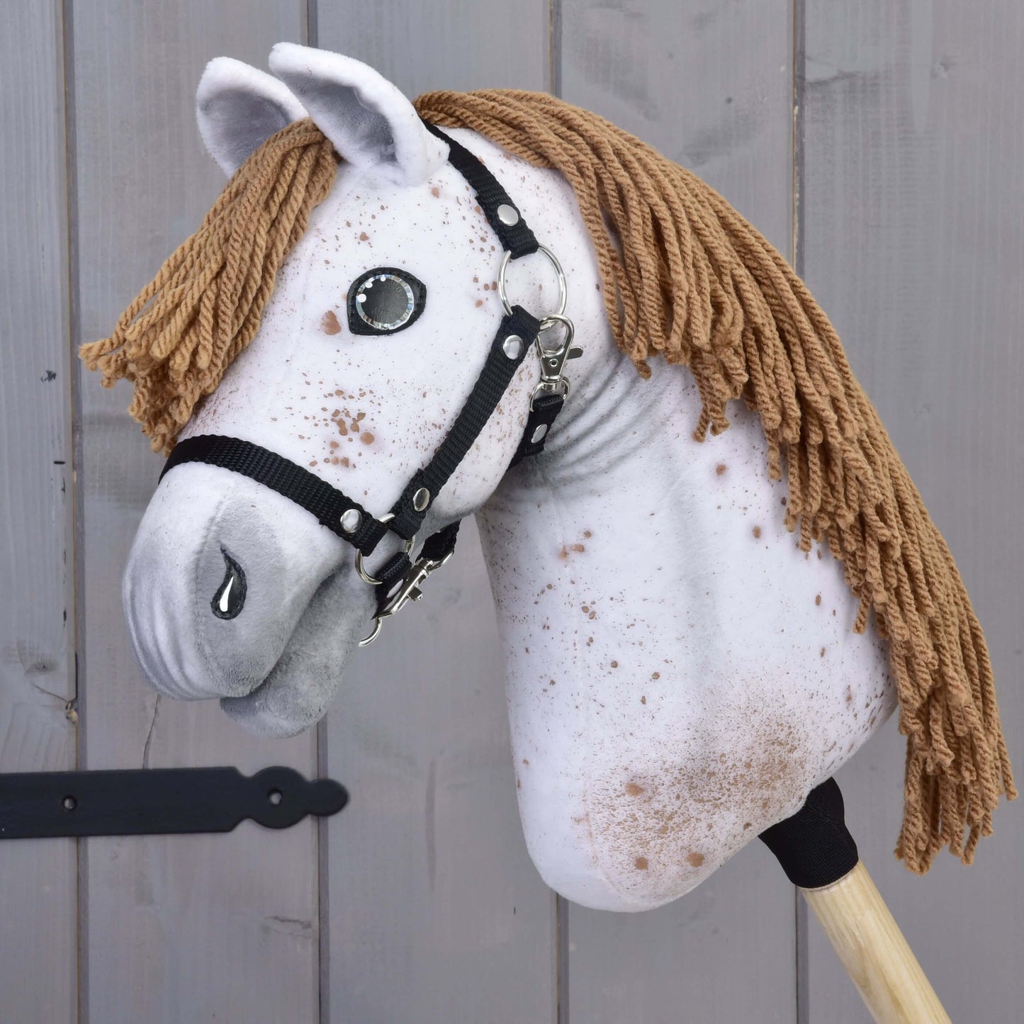 Hobby Horse Rufus s černou ohlávkou od Hobby Horse LarDen s ručně vyrobenou bíle skvrnitou látkovou hlavou, hnědou přízí jako hřívou, knoflíkovýma očima a černou ohlávkou na dřevěné tyči, zobrazený na pozadí šedého dřeva.