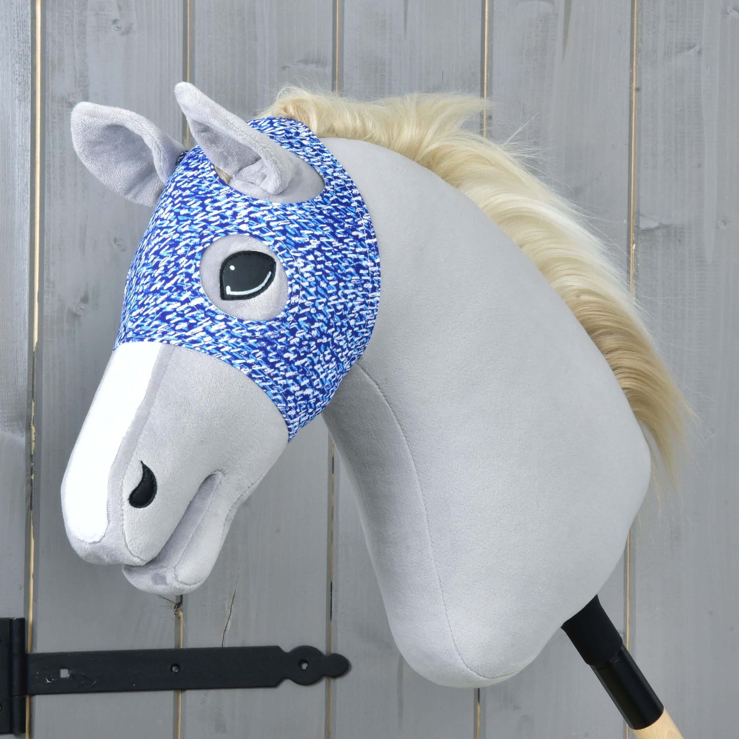 Maska bez uší See od Hobby Horse LarDen se světle šedým obličejem, bílou hřívou, modro-bíle vzorovanou uzdečkou a pratelným obalem na dřevěné tyči, zobrazená před pozadím ze světlých dřevěných panelů.
