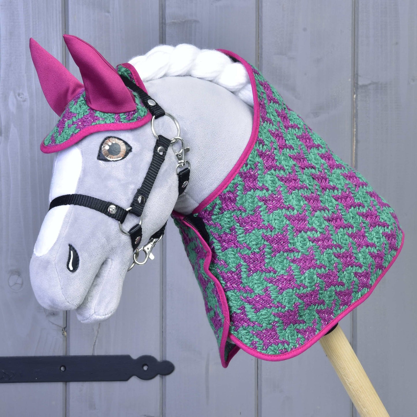 Šedý Hobby Horse LarDen před světle šedou dřevěnou stěnou, na sobě Set Coco: set deky v růžovo-zeleném vzoru kohoutí stopy s ladící růžovou čabrakou na uši a černou uzdečkou.