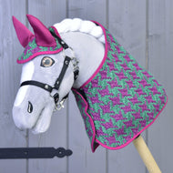 Šedý Hobby Horse LarDen před světle šedou dřevěnou stěnou, na sobě Set Coco: set deky v růžovo-zeleném vzoru kohoutí stopy s ladící růžovou čabrakou na uši a černou uzdečkou.