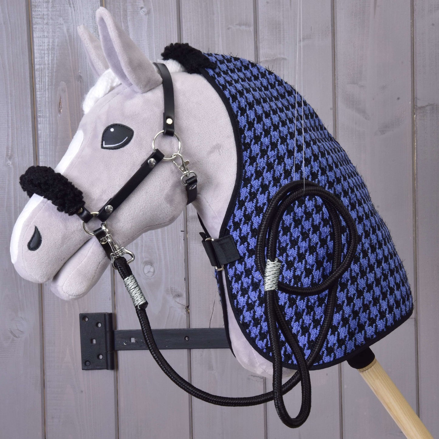 Plyšový Hobby Horse Set Harmony Blue od Hobby Horse LarDen s černou ohlávkou, otěžemi a modro-černou stájovou dekou se vzorem kohoutí stopy je připevněný na stěně na pozadí světlých dřevěných prken.