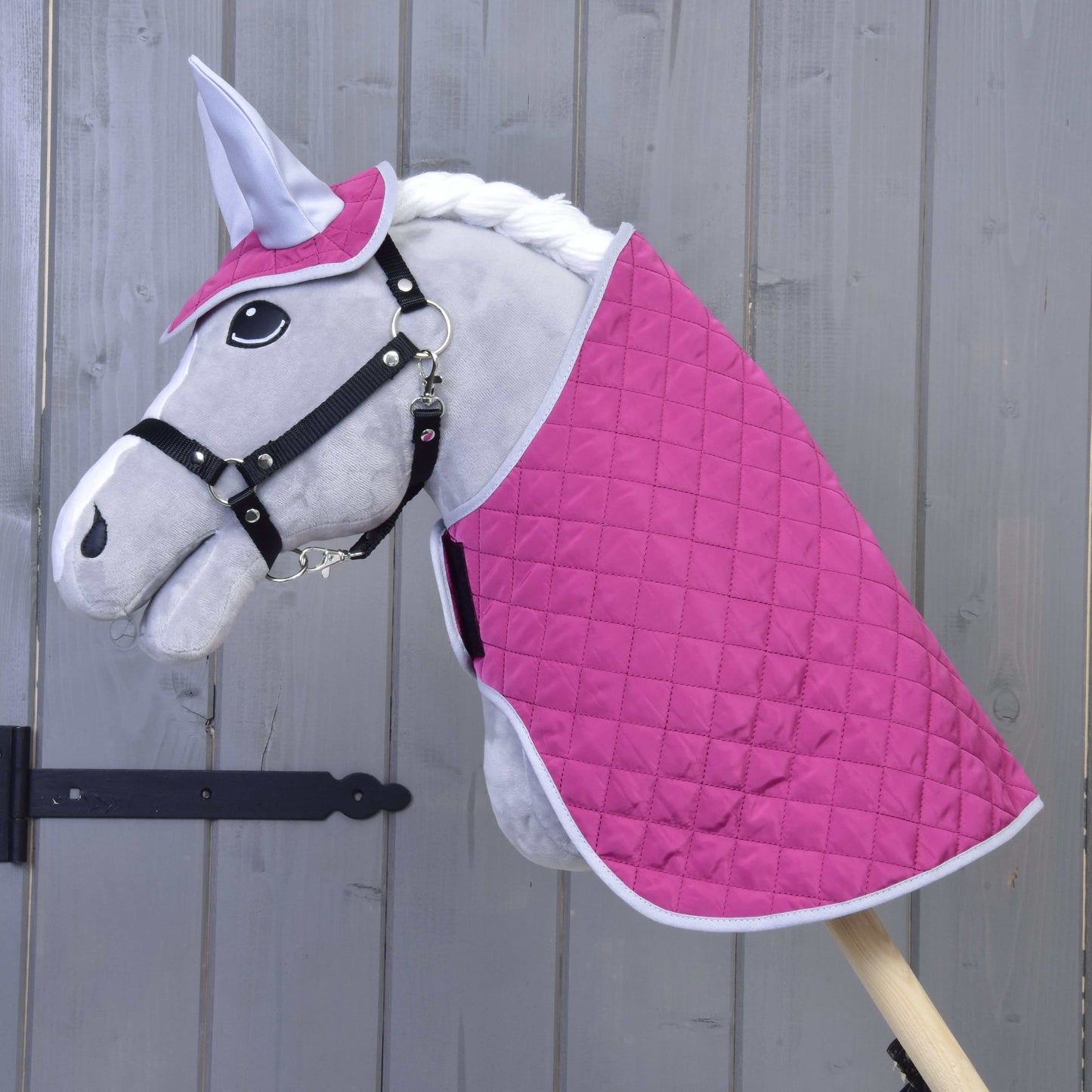 Set Hot Pink Silver od Hobby Horse LarDen: plyšový Hobby Horse se šedým obličejem, černou uzdečkou, bílou hřívou, prošívaným setem deky v sytě růžové a bílé barvě a ladící čabrakou na uši, vše na dřevěné tyči před dřevěnou stěnou.
