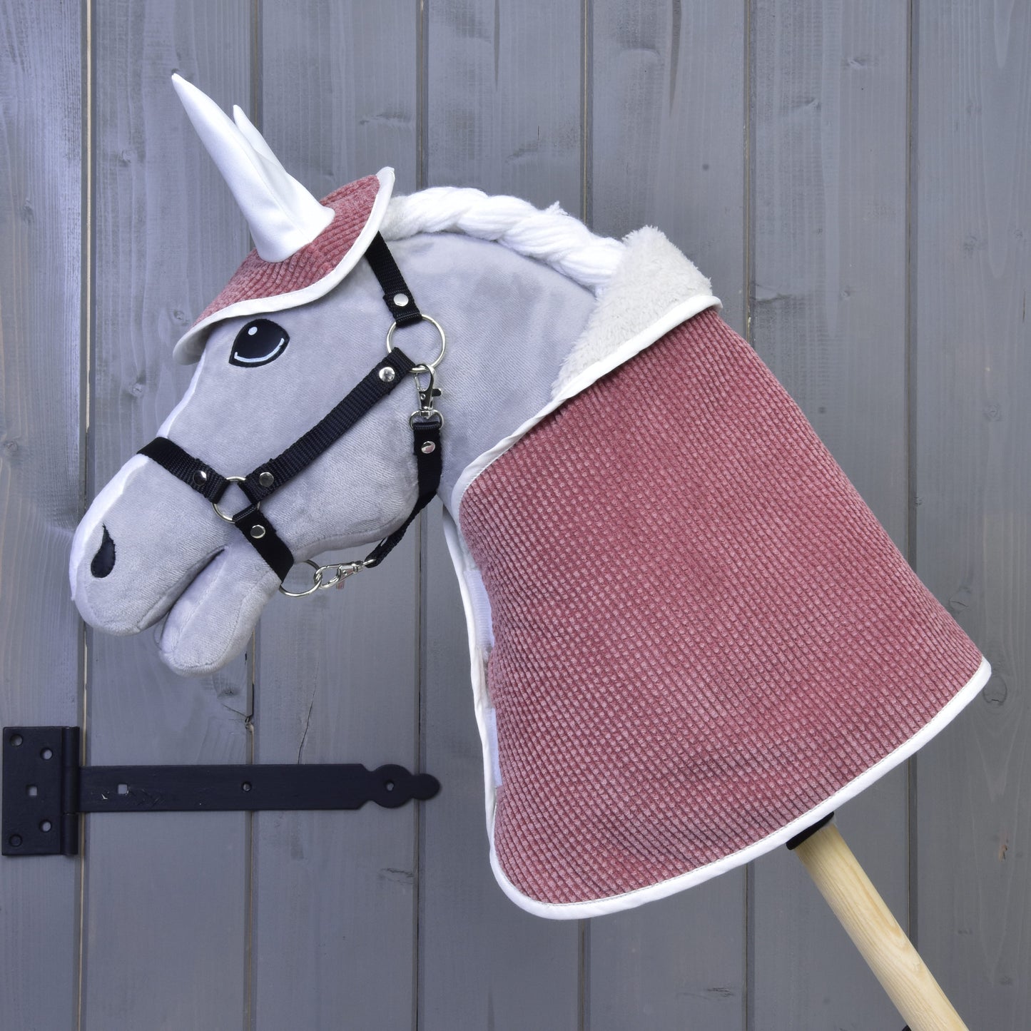 Set Manchester Tea Rose od Hobby Horse LarDen: plyšový jednorožec s bílým rohem, černou uzdečkou, růžovočervenou pokrývkou, čabrakou na uši a bílou hřívou na dřevěné tyči, před šedou dřevěnou stěnou.