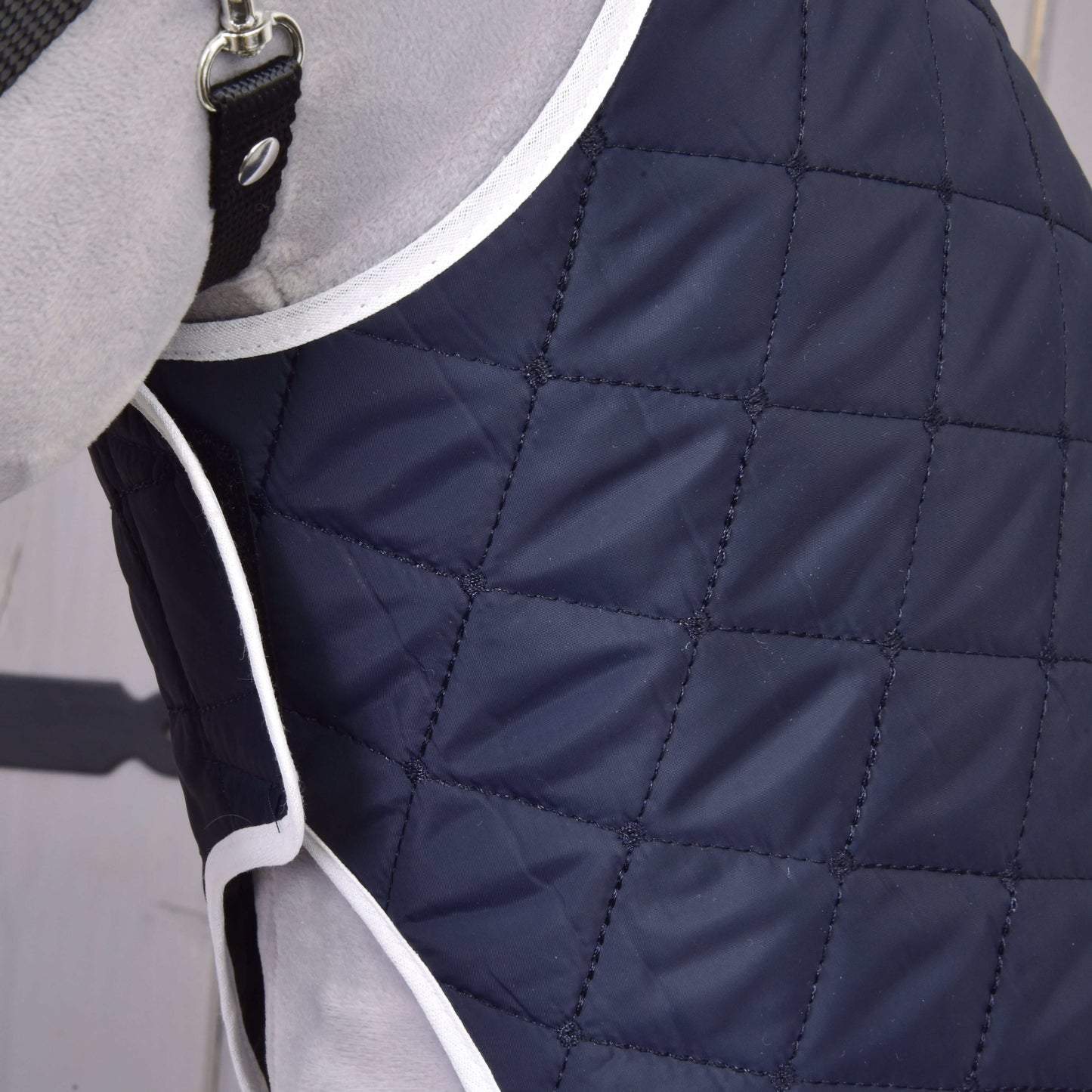 Detail světle šedého psa v prošívaném tmavě modrém kabátku Set Monte od Hobby Horse LarDen s bílým lemováním, kosočtvercovým vzorem a černým páskem s kovovou přezkou pro stylový vzhled deky.