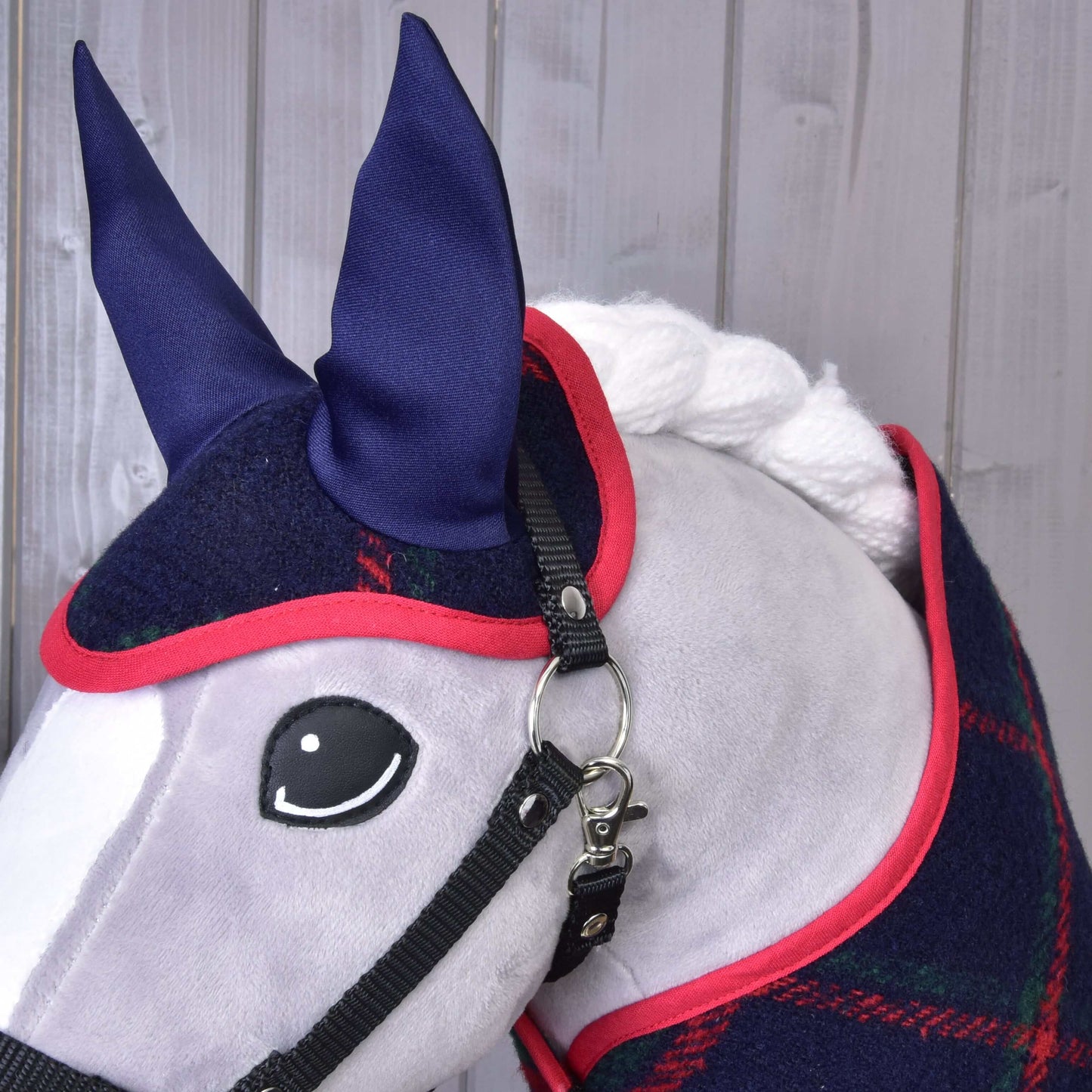 Detail hračkového koně s dekou Set Tartan a ladící čabrakou od Hobby Horse LarDen, s tmavě modrým a červeným kostkováním a modrýma ušima. Kůň má bílou hřívu, černou uzdečku a je před šedým dřevěným pozadím.