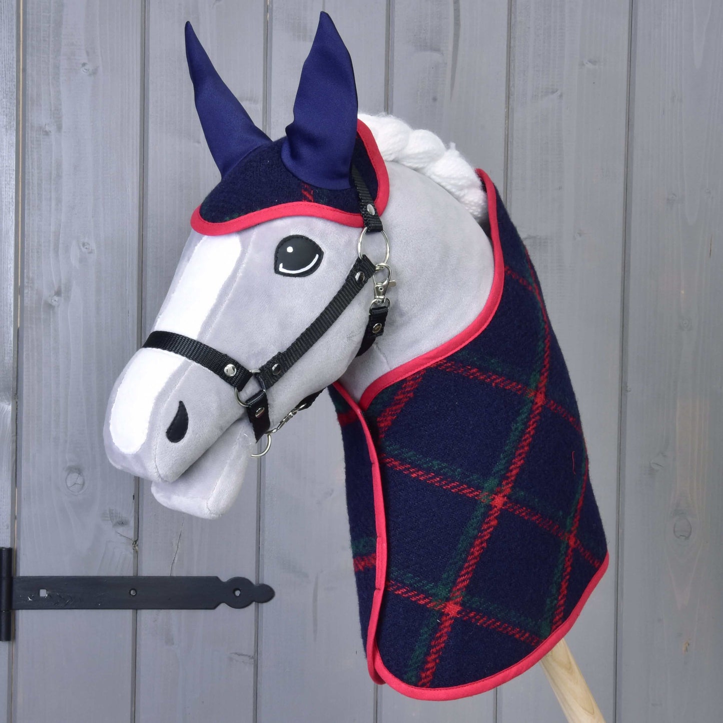 Hobby Horse LarDen „Set Tartan“ s plyšovým Hobby Horse se šedým obličejem, černou uzdečkou, modrýma ušima a tmavě modrou kostkovanou dekou s červeným lemem, před šedou dřevěnou stěnou.
