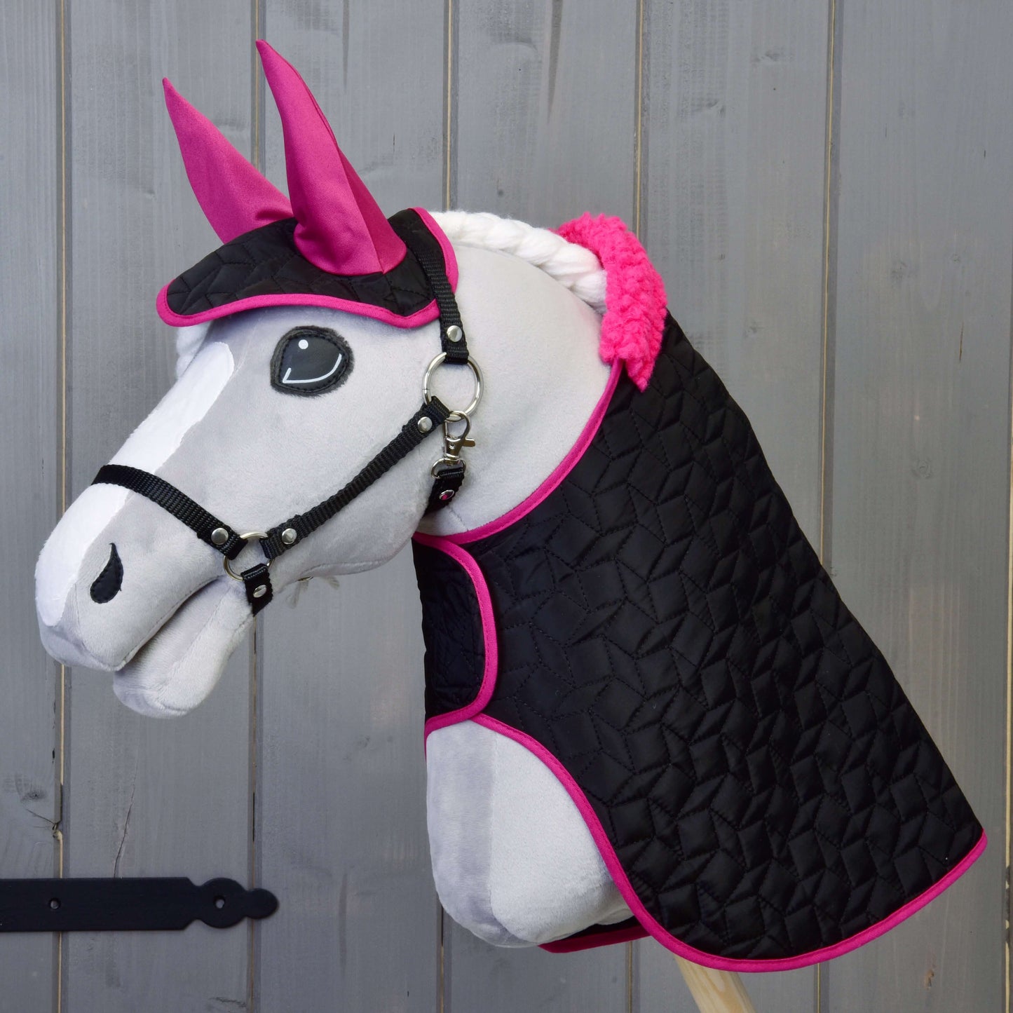 Hobby Horse LarDen "Set Thermo černý s kožíškem" s plyšovým Hobby Horse s bílým obličejem, růžovými ušima, černou uzdečkou a spletenou bílou hřívou, na dřevěném pozadí. Má černou prošívanou deku lemovanou jasně růžovou barvou.