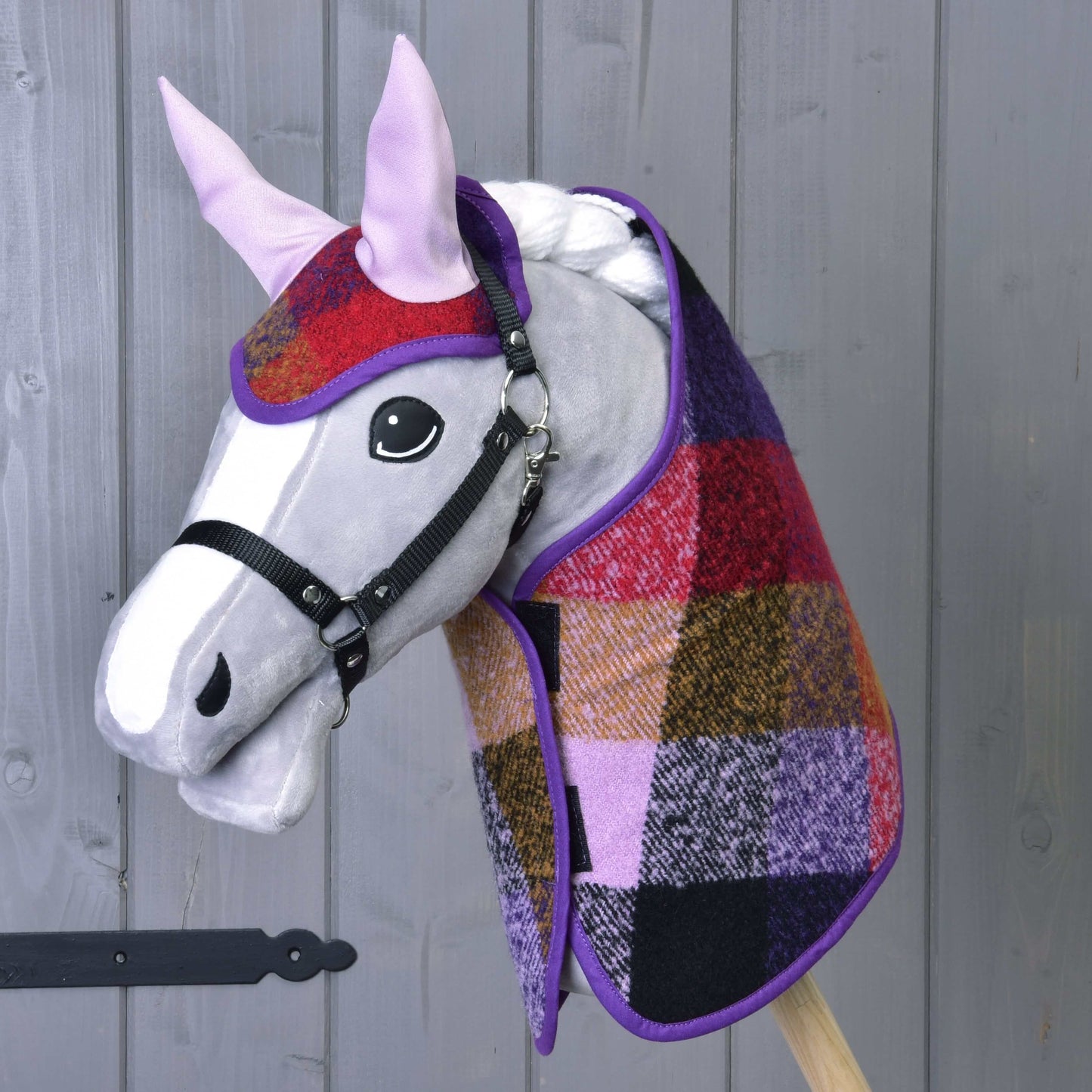 Set podzimní Karo 2 od Hobby Horse LarDen, šedý hobby horse s velkýma černýma očima, oblečený do červeno-fialovo-žluto-černé kostkované deky a ladící čabraky na dřevěné tyči před šedou stěnou.