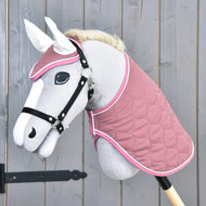 Hobby Horse LarDen „Set Dusty Rose“ na šedém Hobby Horse s bílou hřívou, černou uzdečkou, čabrakou, prošívanou růžovou dekou a černou tyčí, zobrazeném před dřevěnou stěnou.