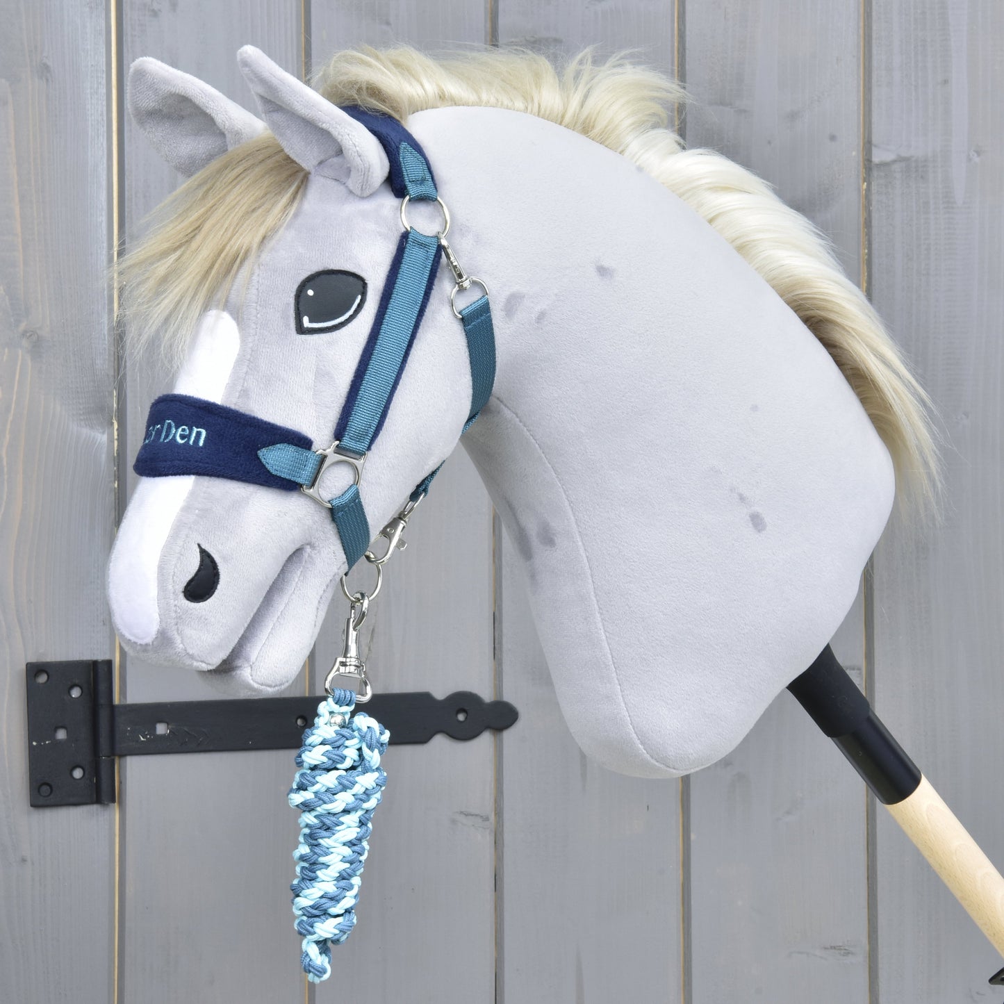 Set LarDen Exclusive Blue od Hobby Horse LarDen, šedý Hobby Horse s modrou ohlávkou a krouceným modrobílým vodítkem, nasazený na dřevěné tyči, na pozadí dřevěného plotu.