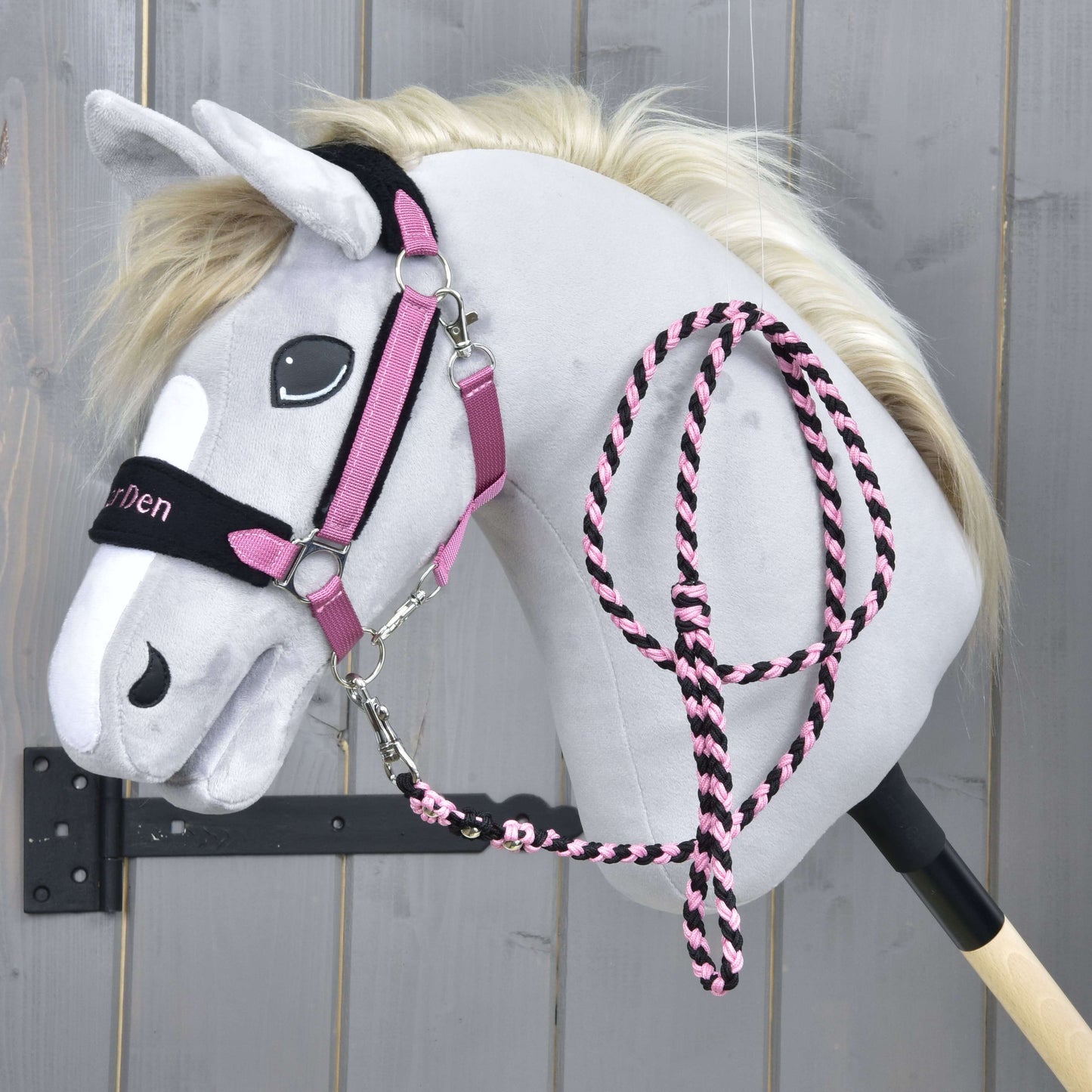Set LarDen Exclusive Pink od Hobby Horse LarDen, šedý plyšový Hobby Horse s krémovou hřívou, růžovo-černou uzdečkou, ladícím fleecovým nánosníkem a vodítkem na černé tyči, na dřevěném pozadí.
