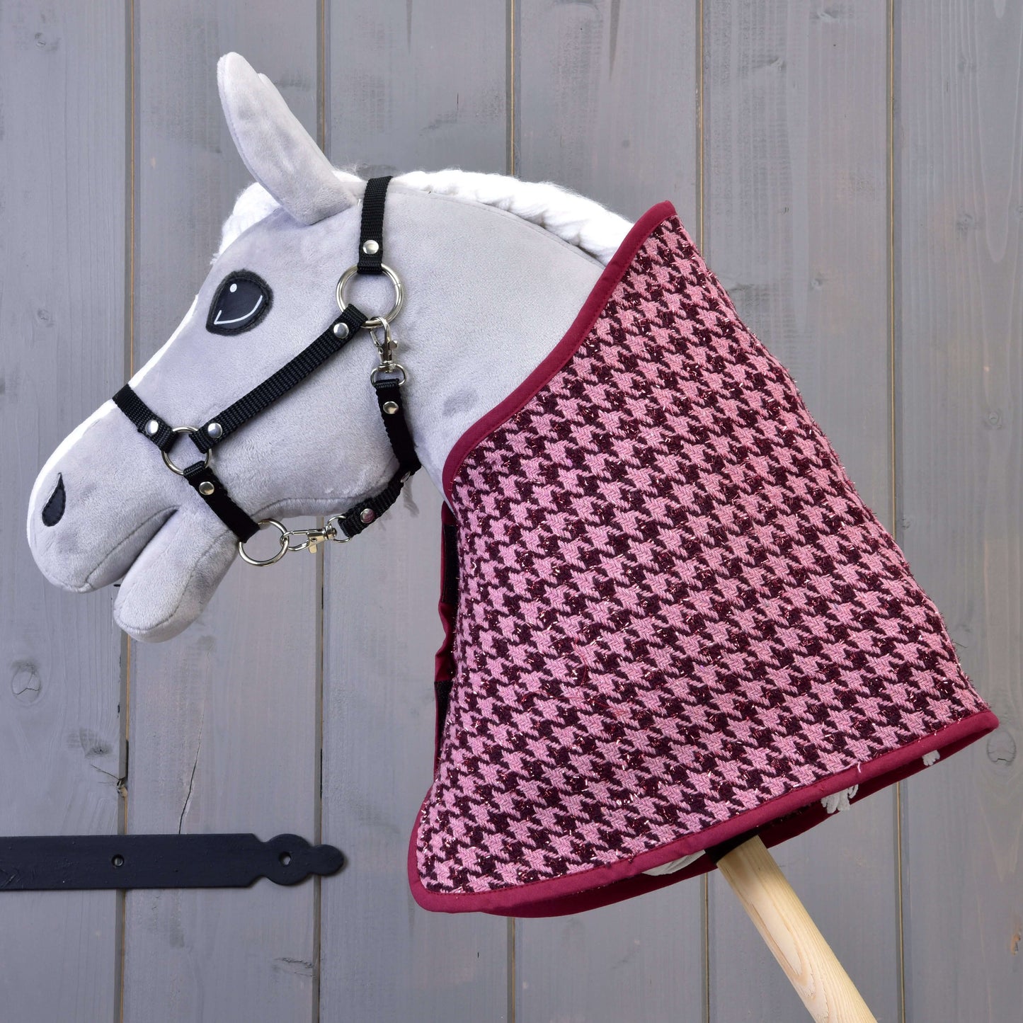 Hobby Horse LarDen "Set slavnostní Bordeaux" s šedým plyšovým Hobby Horse s černou uzdečkou a vínovo-růžovou dekou se vzorem kohoutí stopy, nasazeným na dřevěné tyči na pozadí šedých dřevěných prken.