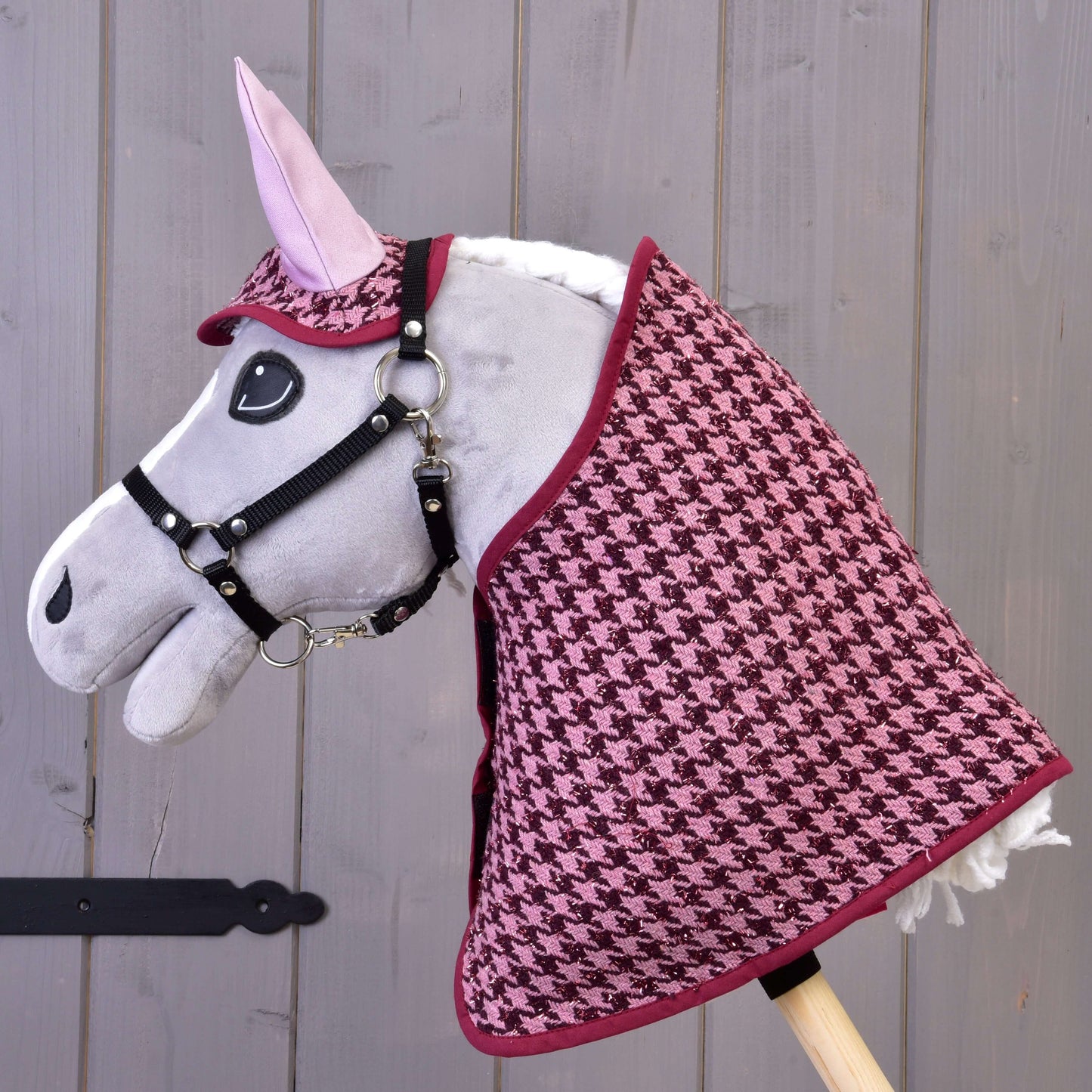Šedá hračka koně na tyči od Hobby Horse LarDen má na sobě růžovo-černou deku se vzorem kohoutí stopy a set s čabrakou Set slavnostní Bordeaux, ideální jako dekorace koně, aranžovaná před šedou dřevěnou stěnou.
