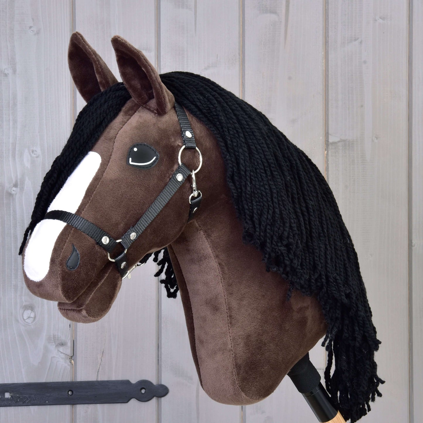 Hobby Horse Sisi s černou ohlávkou od Hobby Horse LarDen, s tmavě hnědou srstí, bílým pruhem na hlavě, černou přízovou hřívou a vzhledem mladého hanoverského sportovního koně, na pozadí dřevěných panelů.