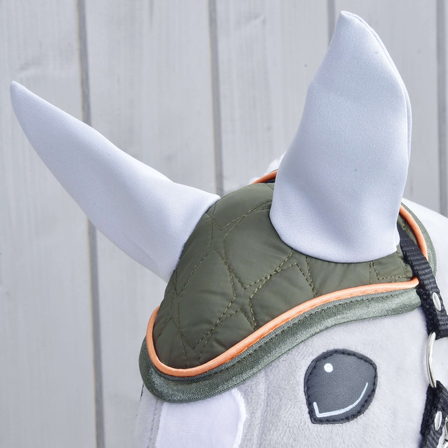 Detail plyšového Hobby Horse LarDen s čabrakou Sport, zelenou prošívanou čabrakou s oranžovým lemováním a měkkými vzpřímenými světle šedými ušima, na světlém dřevěném pozadí.