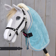 Plyšový Hobby Horse od Hobby Horse LarDen se světle šedým obličejem, černou uzdečkou, bílou tlamou a dekou Deka stájová Fern ze světle modrého materiálu PES, pózující na dřevěném pozadí.