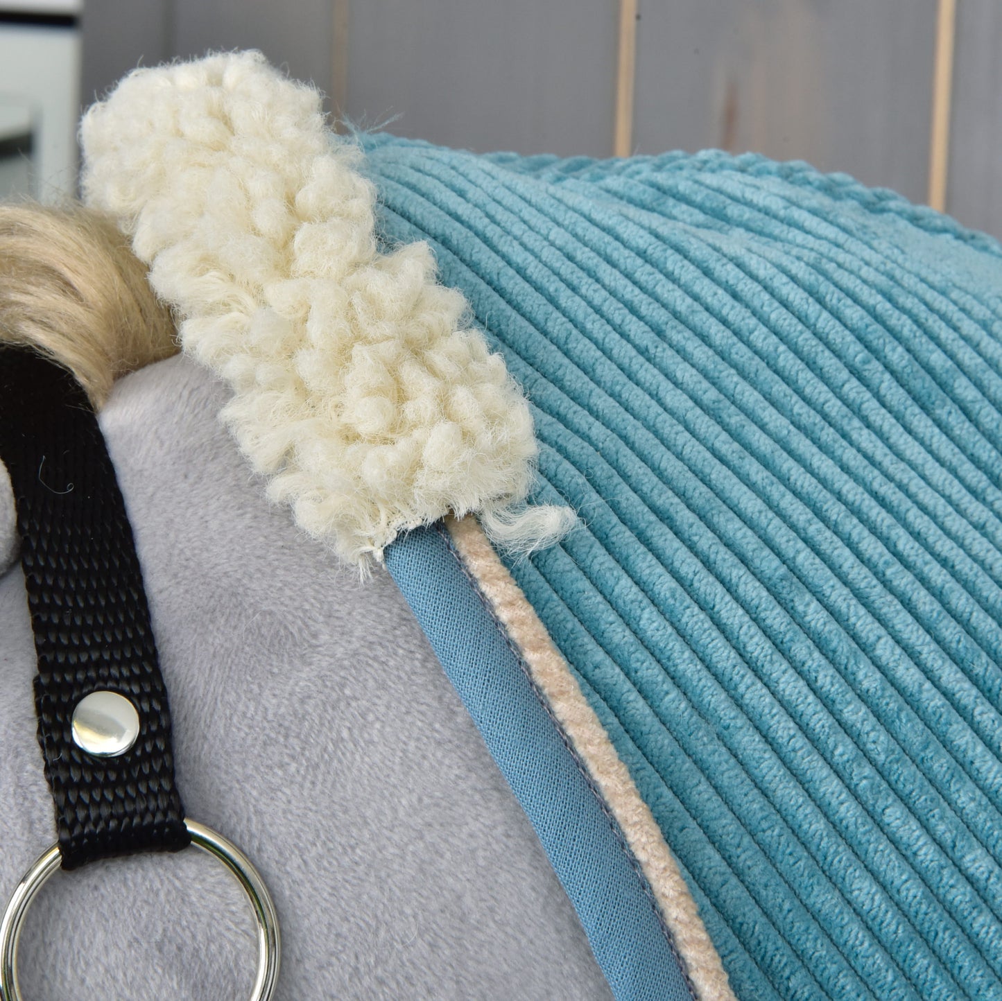 Detail světle šedého plyšového koně s modrou dekou Deka stájová Fern od Hobby Horse LarDen, s krémovým fleecovým lemováním a černým páskem se stříbrným kroužkem. V pozadí jsou vidět světlé dřevěné panely.