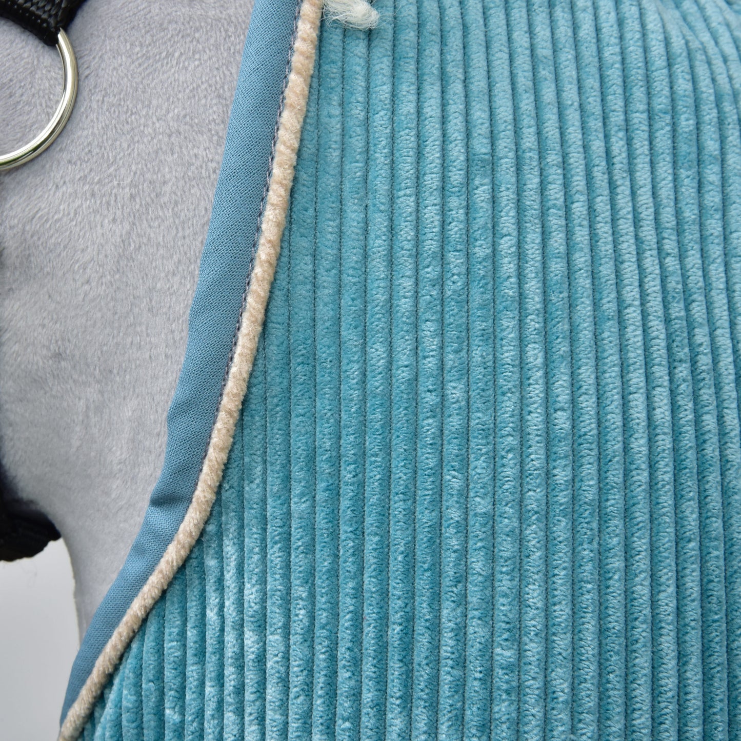Detail texturované modré deky Deka stájová Fern od Hobby Horse LarDen přehozené přes šedého koně, s béžovým chlupatým lemem a viditelným kovovým kroužkem z koňského postroje.