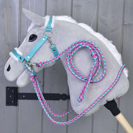 Set Perfect Combination Turquoise od Hobby Horse LarDen, plyšový šedý Hobby Horse s bílou hřívou, tyrkysovou uzdečkou, ohlávkou s kožešinovým polstrováním a růžovo-modrými provazovými otěžemi, visí před šedou dřevěnou stěnou.