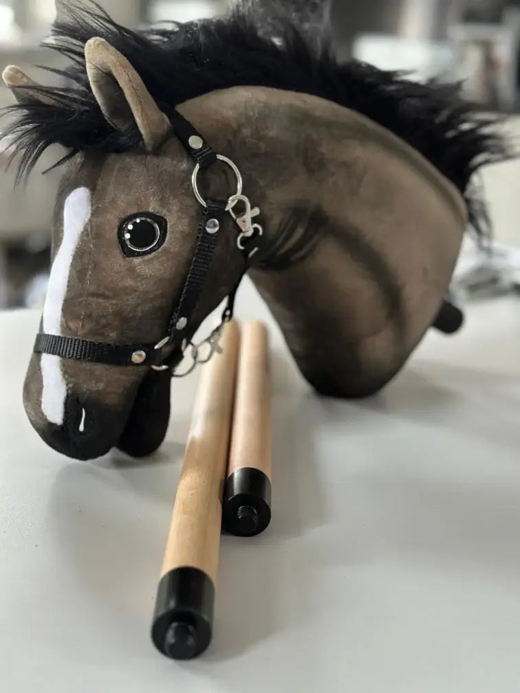 Plyšová hlava Hobby Horse s černou hřívou a ohlávkou leží na stole vedle dvou dřevěných tyčí s černými koncovkami, ideální pro nadšence do Hobby Horsing.