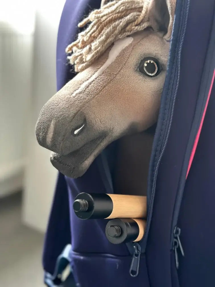 Plyšový Hobby Horse se světlou hřívou a bílou lysinou vykukuje z částečně rozepnuté tmavě modré tašky, uvnitř tašky jsou vidět také dvě dřevěné tyče s držadly — ideální pro dobrodružství při Hobby Horsing.