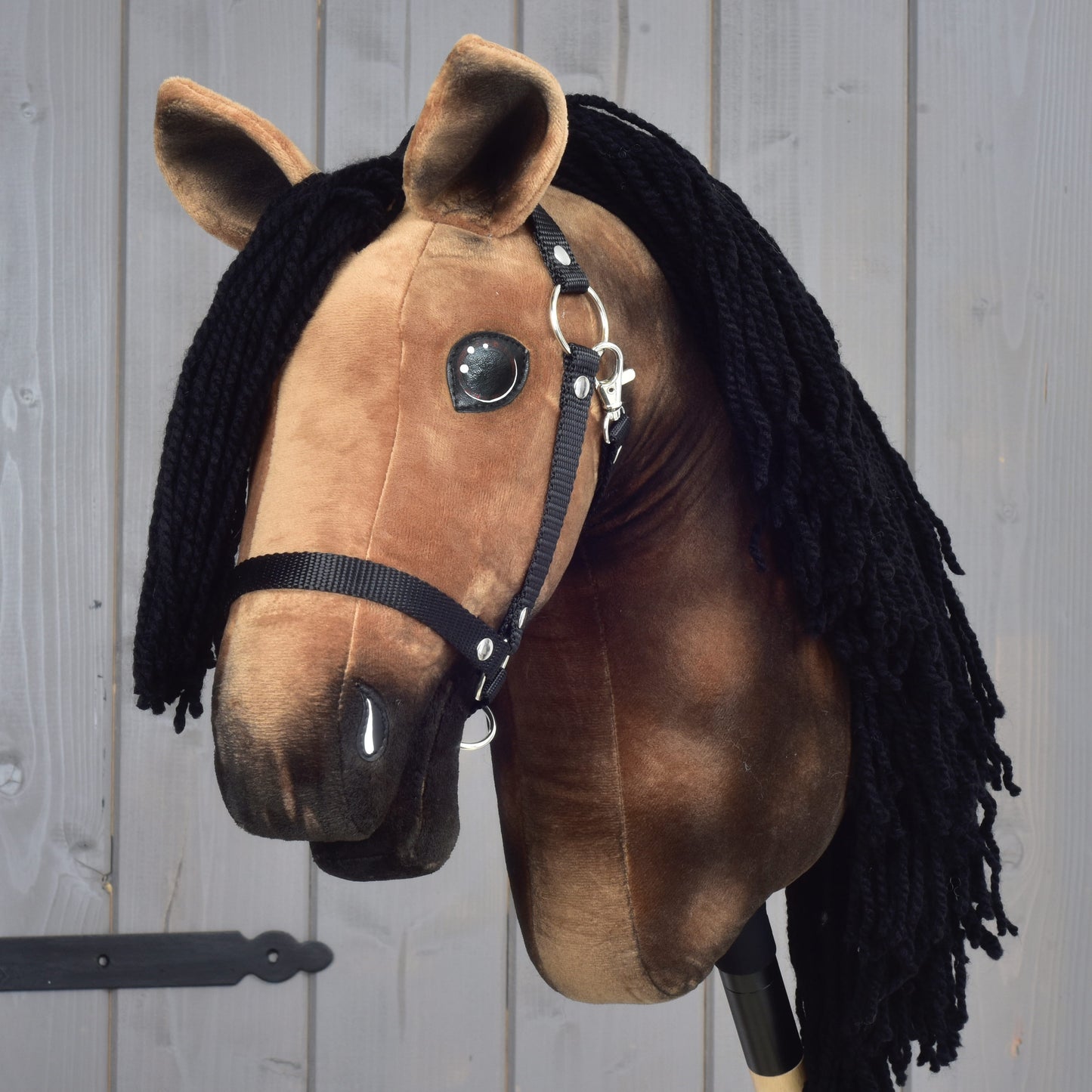 Hobby Horse Stormy s černou ohlávkou od Hobby Horse LarDen, s plyšovým hnědým tělem a dlouhou černou přízovou hřívou, před světle šedou dřevěnou stěnou.