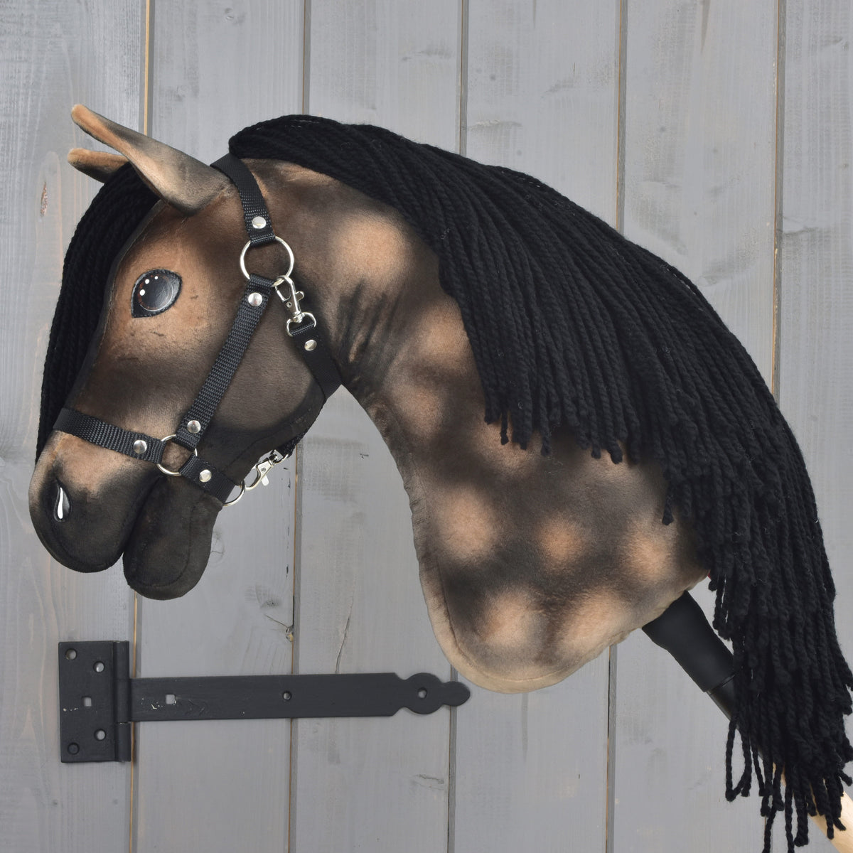 Hobby Horse Stormy s černou ohlávkou od Hobby Horse LarDen, realistický model inspirovaný hannoverským hřebcem s černou přízovou hřívou, připevněný na šedé dřevěné stěně černým držákem.