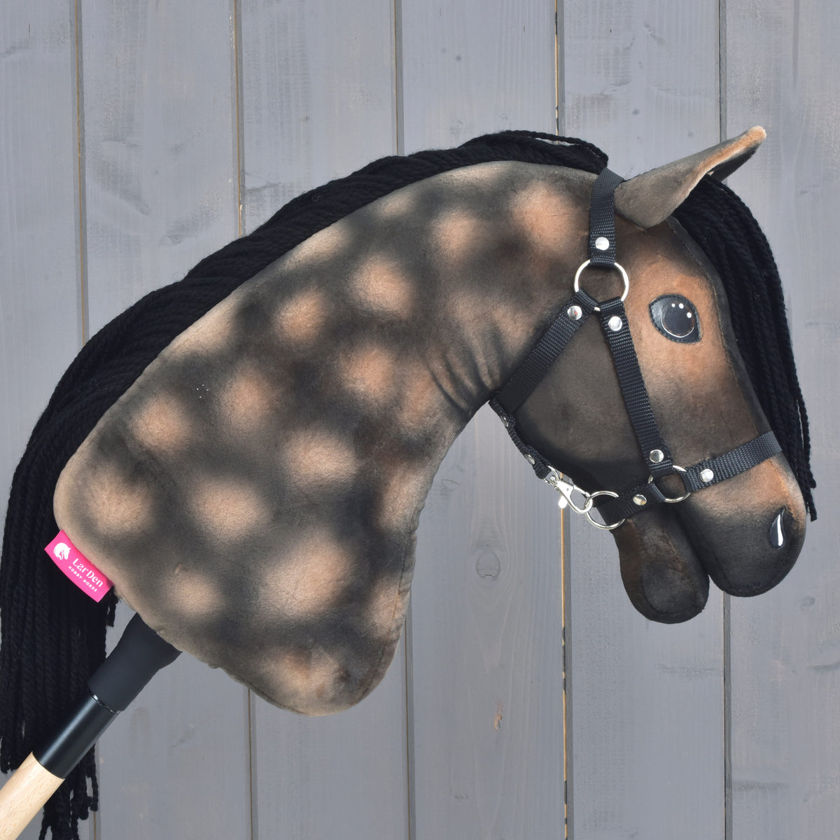 Hobby Horse Stormy s černou ohlávkou od Hobby Horse LarDen s grošovanou hnědo-černou plyšovou hlavou inspirovanou hannoverským hřebcem, doplněný černou přízovou hřívou a černou ohlávkou, na světlém dřevěném pozadí.