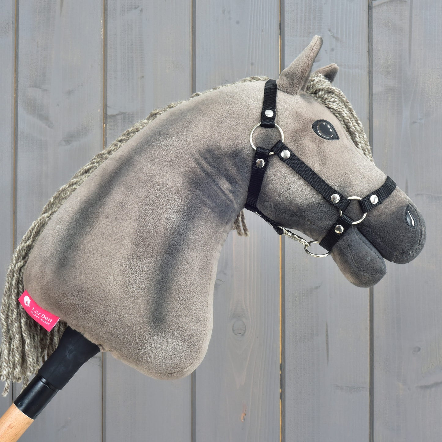 Hobby Horse Swing s černou ohlávkou - velikost S od Hobby Horse LarDen, s plyšovým provedením, šedou hřívou a na dřevěné tyči, zachycující vznešený půvab hanoverského hřebce, vystavený před oprýskaným šedým dřevěným pozadím.