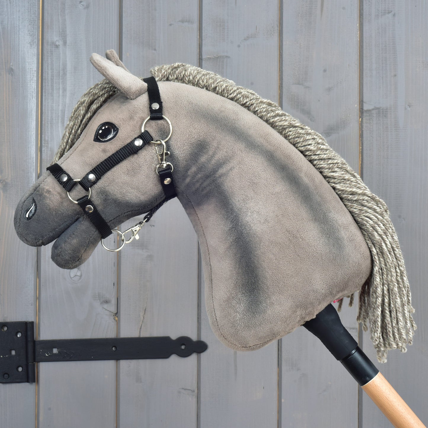Hobby Horse Swing s černou ohlávkou - velikost S od Hobby Horse LarDen, šedý plyšový Hobby Horse připomínající hanoverského hřebce, s černou ohlávkou a provazovou hřívou, na dřevěné tyči, vystavený před dřevěnou stěnou s viditelnou kresbou dřeva a černým kovovým závěsem.