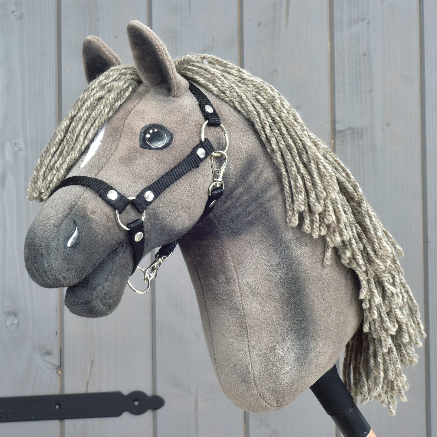 Hobby Horse LarDen "Hobby Horse Swing with black halter", velikost S, plyšový koník se zapletenou přízovou hřívou a elegantní černou ohlávkou, nasazený na tyči a vystavený před dřevěným pozadím.