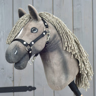 Hobby Horse LarDen "Hobby Horse Swing with black halter", velikost S, plyšový koník se zapletenou přízovou hřívou a elegantní černou ohlávkou, nasazený na tyči a vystavený před dřevěným pozadím.