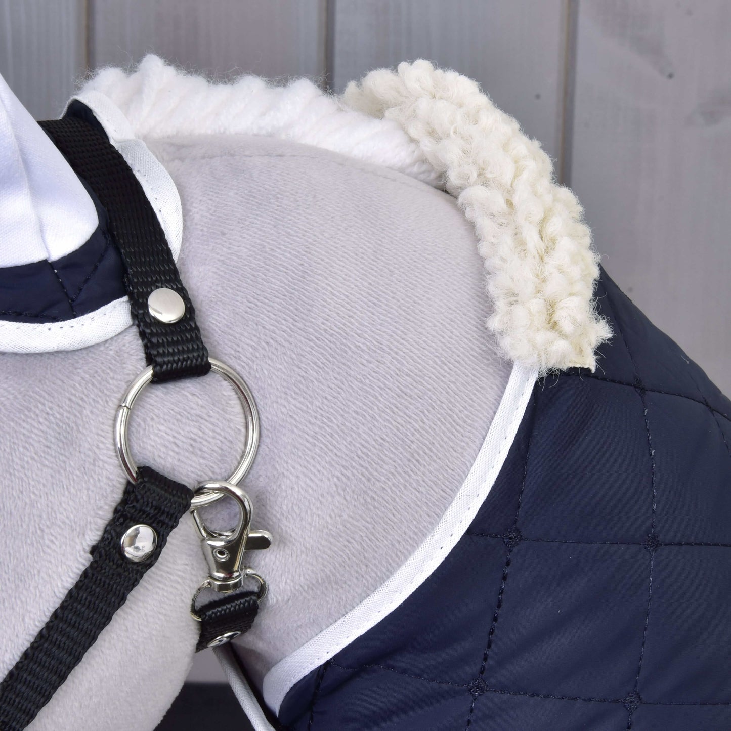 Detail psího ramene v kabátku Set Thermo Blue od Hobby Horse LarDen s prošívanou tmavě modrou látkou, bílým lemováním a huňatým límcem, doplněný černým postrojem s kovovým kroužkem. V pozadí je vidět dřevěné obložení.