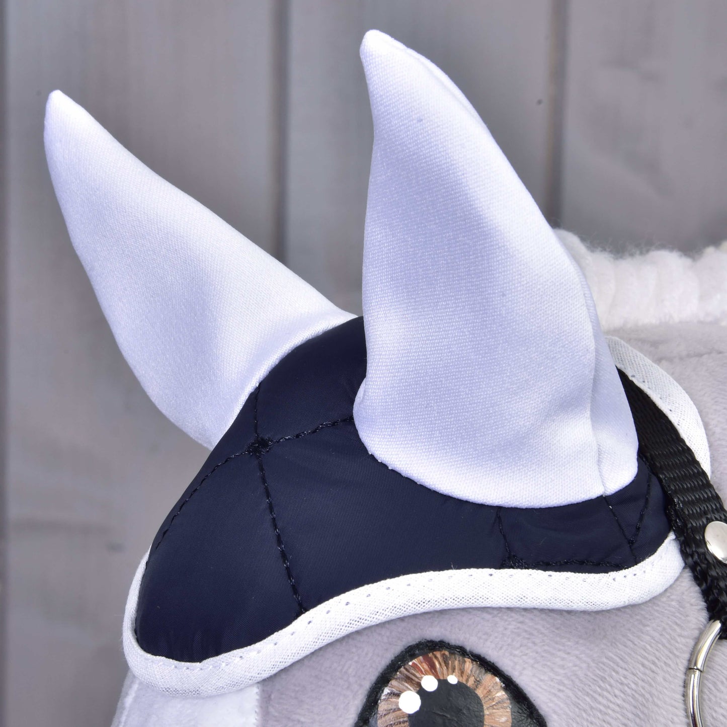 Detail hlavy plyšového koně v elegantním Set Thermo Blue od Hobby Horse LarDen, s bílýma ušima, tmavě modro-bílou čepicí a černou uzdečkou. Perfektní dekorativní kousek na pozadí světlých dřevěných panelů.