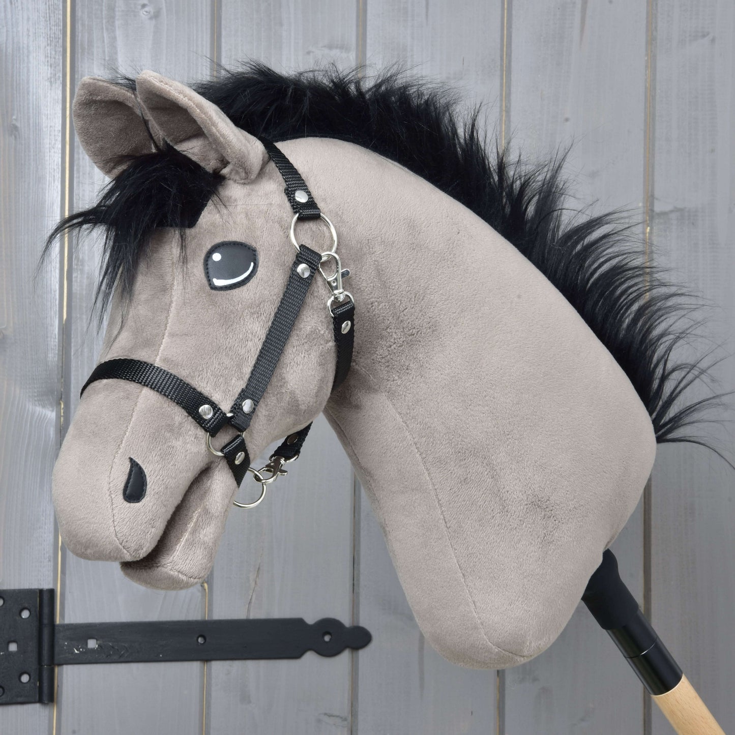 Hobby Horse Thor s černou ohlávkou od Hobby Horse LarDen, plyšový hanoverský hřebec Hobby Horse s černou hřívou, černou ohlávkou a vyšívanýma očima, na dřevěné tyči před světle šedou dřevěnou stěnou.
