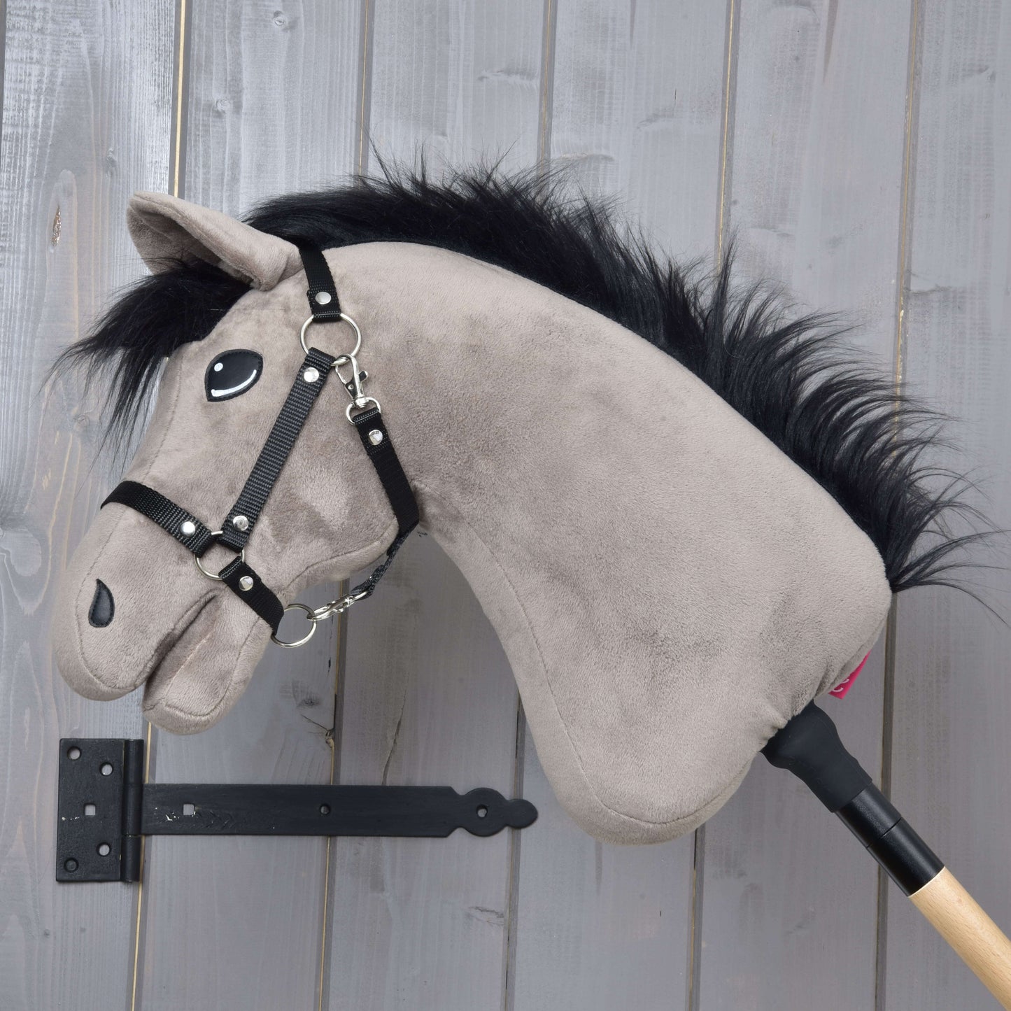 Hobby Horse Thor s černou ohlávkou od Hobby Horse LarDen, se šedou plyšovou hlavou v hanoverském stylu a černou hřívou, na dřevěné tyči, vystavený na šedé stěně pomocí černého kovového držáku.