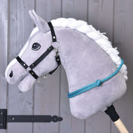 Nákrčník Tyrkys od Hobby Horse LarDen na šedém plyšovém Hobby Horse s bílou přízovou hřívou, černou uzdečkou, modrými provazovými otěžemi a ručně vyrobeným tyrkysovým cordeem pro ježdění bez uzdečky, na tyči před dřevěnou stěnou.