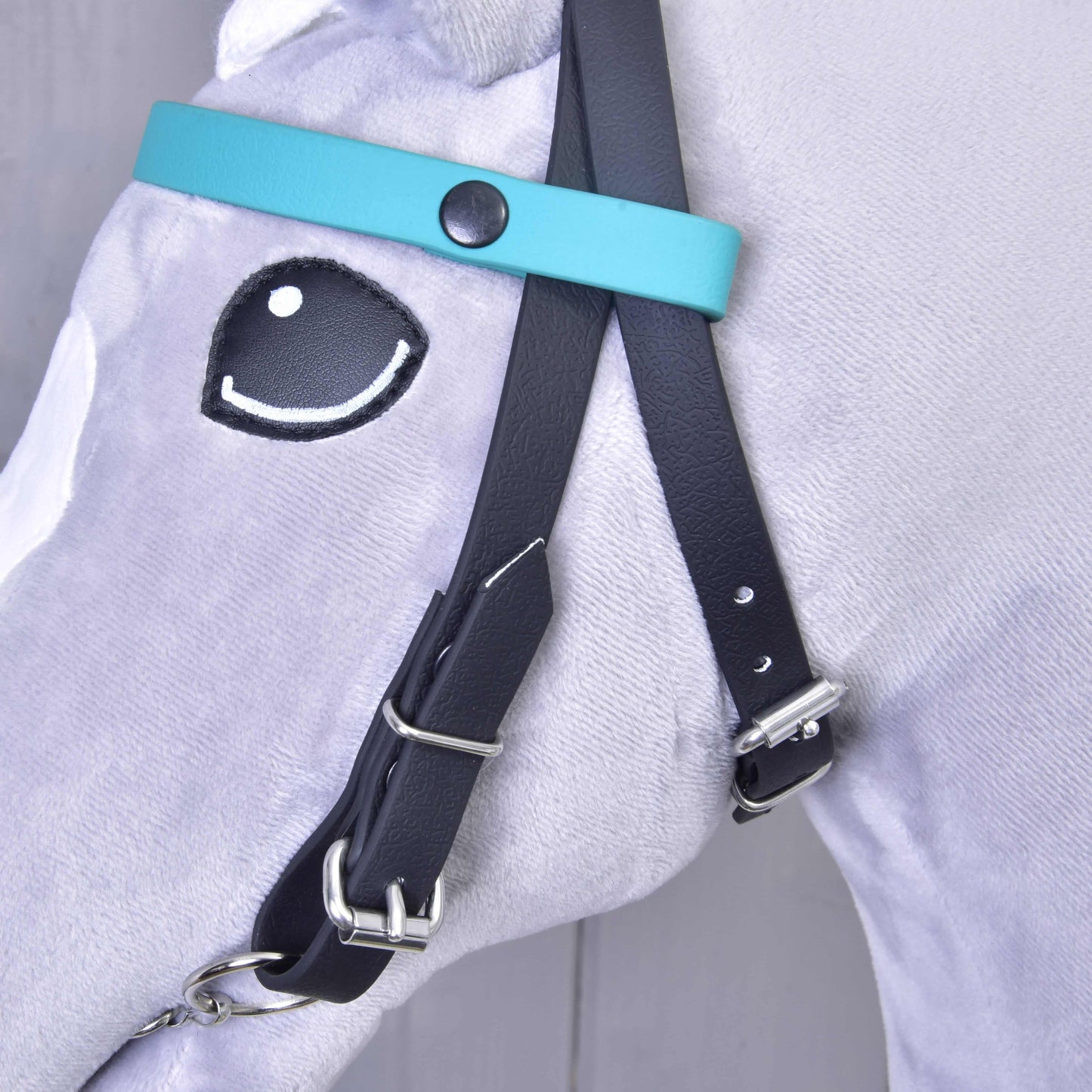 Detail uzdečky bez nánosníku tyrkysová se 2 čelenkami (vel. M) od Hobby Horse LarDen na světle šedém plyšovém koni s vyšívaným okem v kresleném stylu. Uzdečka má stříbrné přezky a vyměnitelnou čelenku, bez nánosníku, v tyrkysové a černé barvě.