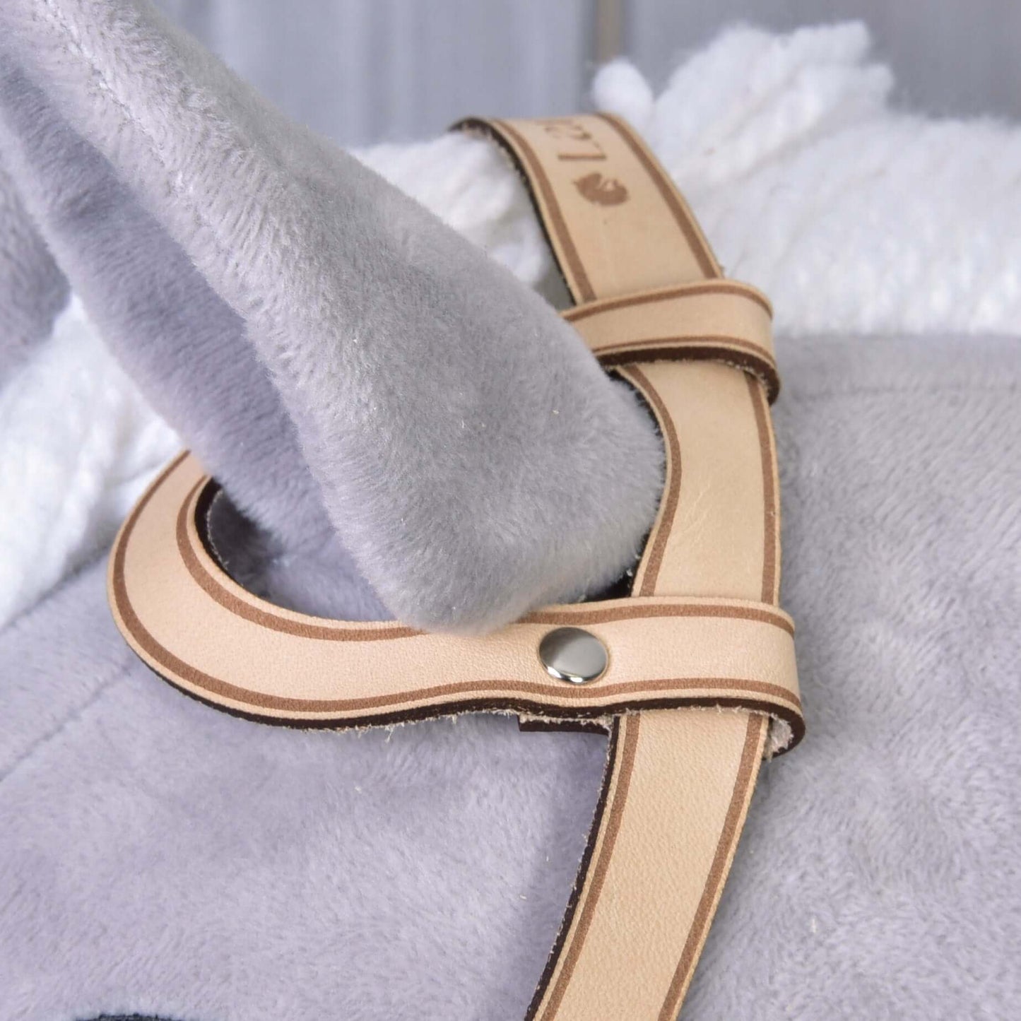 Detail šedé plyšové látky a měkké bílé deky s uzdečkou western jednouška od Hobby Horse LarDen – světle hnědým řemínkem z kvalitní kůže s kovovými nýty, provlečeným kolem svěšeného šedého plyšového ucha.