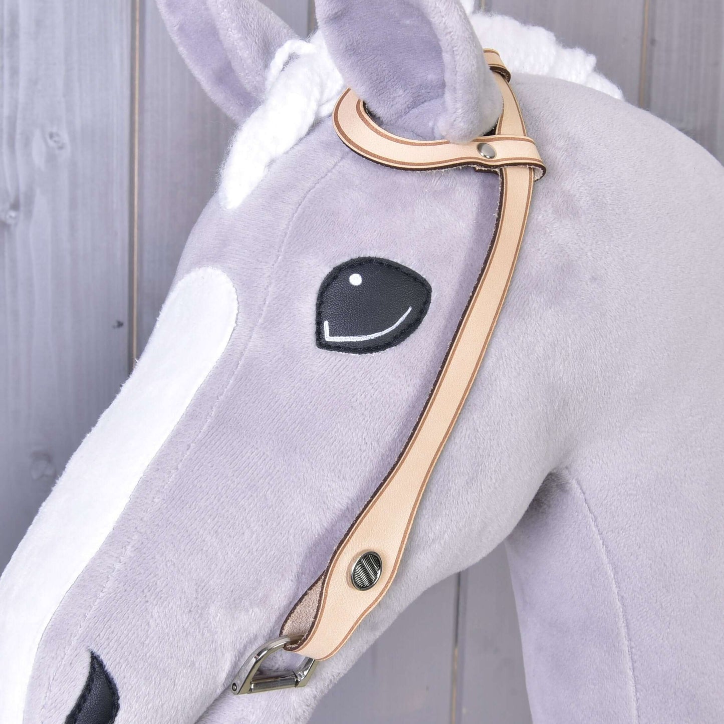 Detail hlavy plyšového koně ze světle šedé a bílé látky s vyšívaným černým okem a uzdečkou western jednouška od Hobby Horse LarDen se stříbrným kováním na dřevěném pozadí.