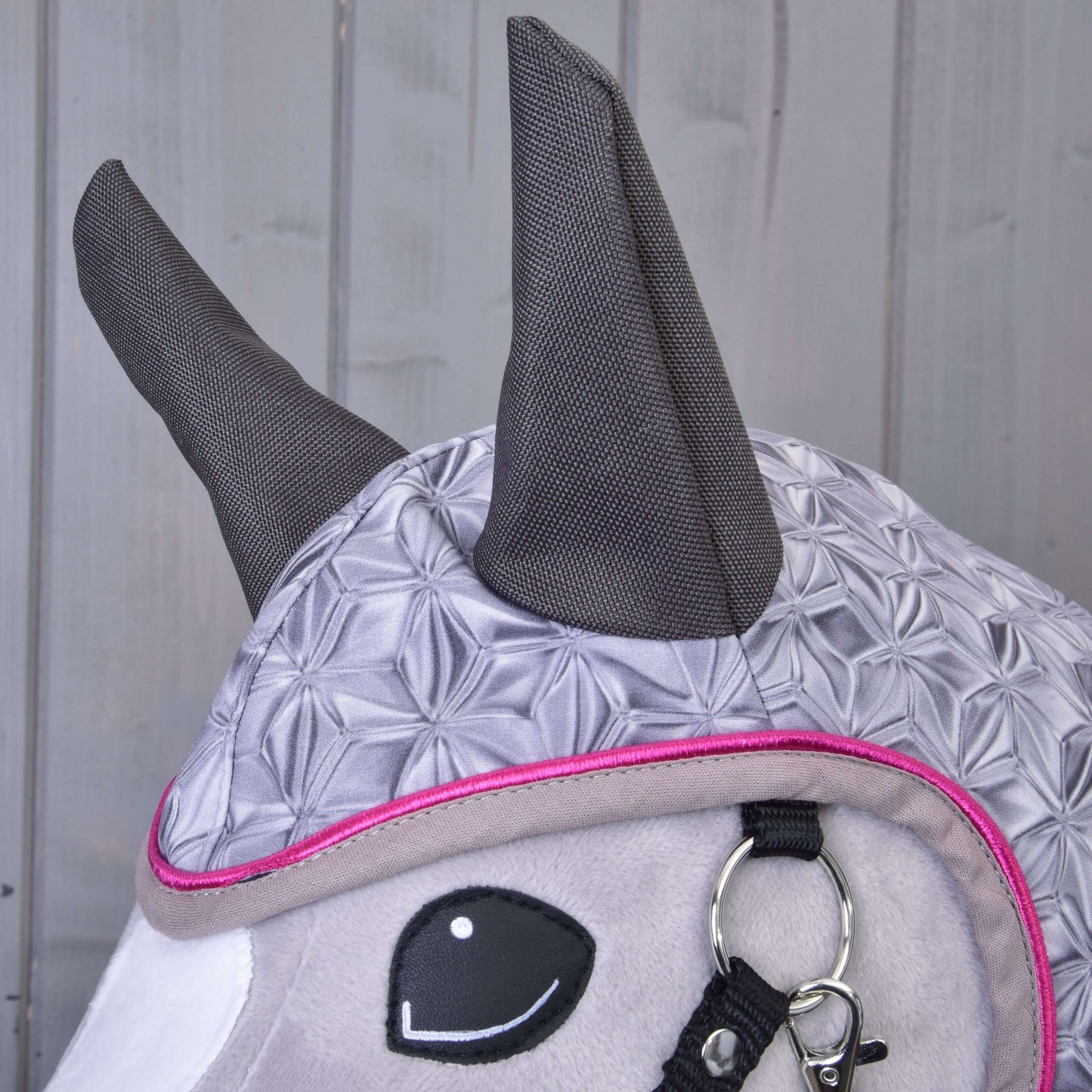Detail plyšové koňské hračky s Hobby Horse LarDen Pláštěnka Valentine s šedým vzorovaným designem, tmavě šedýma ušima, růžovým lemováním a černo-stříbrnou uzdečkou na dřevěném pozadí.