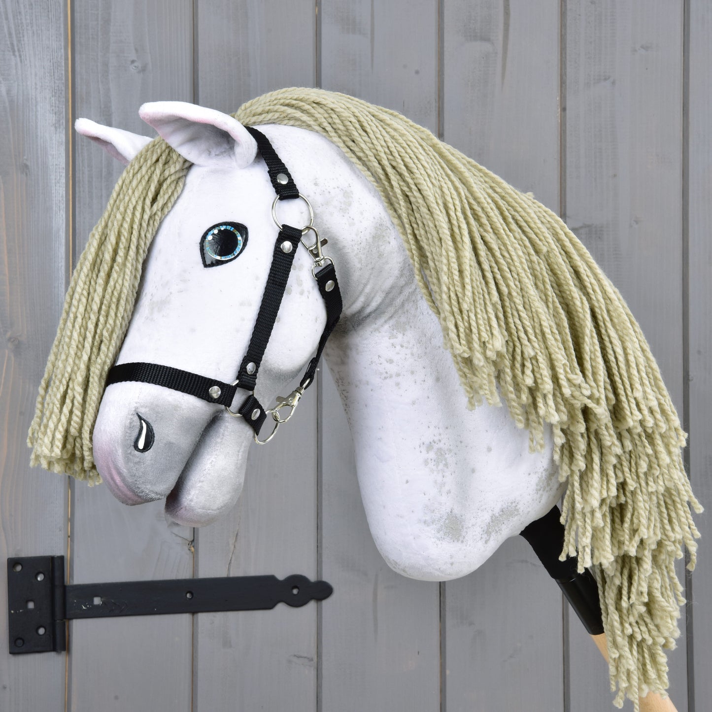 Hobby Horse Victory s černou ohlávkou - velikost M od Hobby Horse LarDen, s bílou plyšovou hlavou, blond hřívou z příze, černou ohlávkou a malovaným modrým okem, na dřevěné tyči, před šedou dřevěnou stěnou.