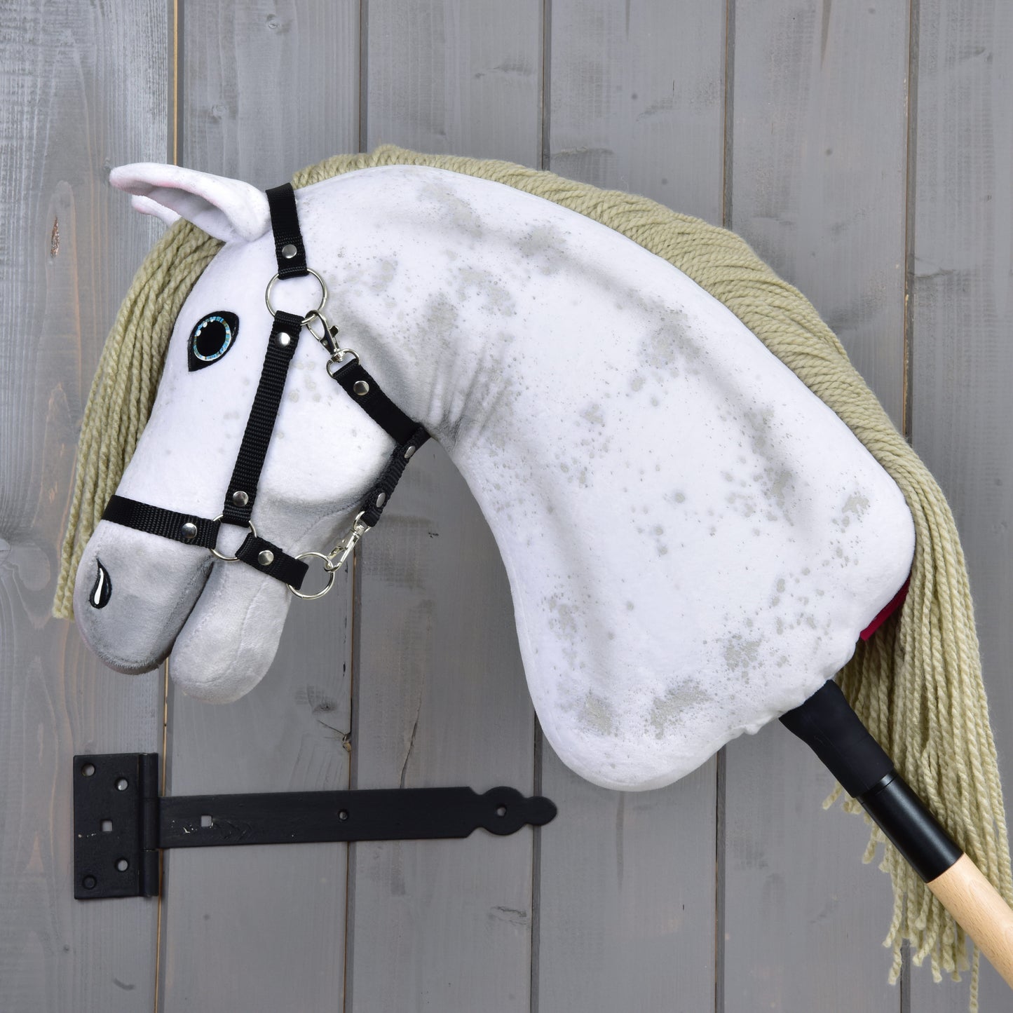 Hobby Horse Victory s černou ohlávkou - velikost M od Hobby Horse LarDen, s bílým plyšovým provedením, dlouhou hřívou z příze a černou ohlávkou, vystavený na šedé dřevěné stěně s černou kovovou konzolí.