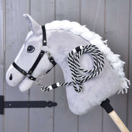 Hobby Horse LarDen „Hobby Horse - vodítko proužek černé“ zobrazuje plyšového koně s bílou hřívou, černou uzdečkou a černobíle pruhovaným vodítkem na dřevěné tyči, vystaveného před dřevěnou stěnou.
