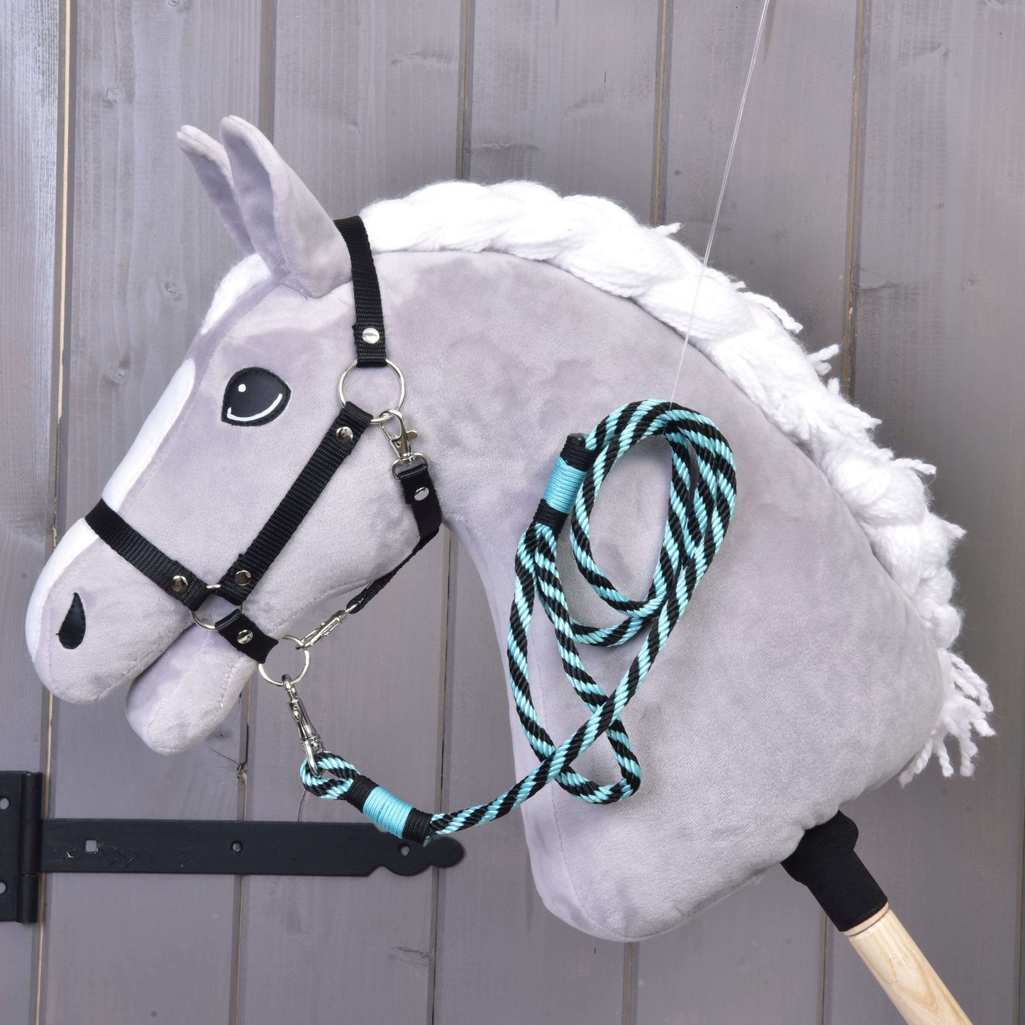 Hobby Horse LarDen „Hobby Horse - vodítko proužek zelené“ zobrazuje plyšového šedého koně s bílou hřívou, černou uzdečkou a zeleným pruhovaným vodítkem, nasazeného na dřevěné tyči před dřevěnou stěnou.