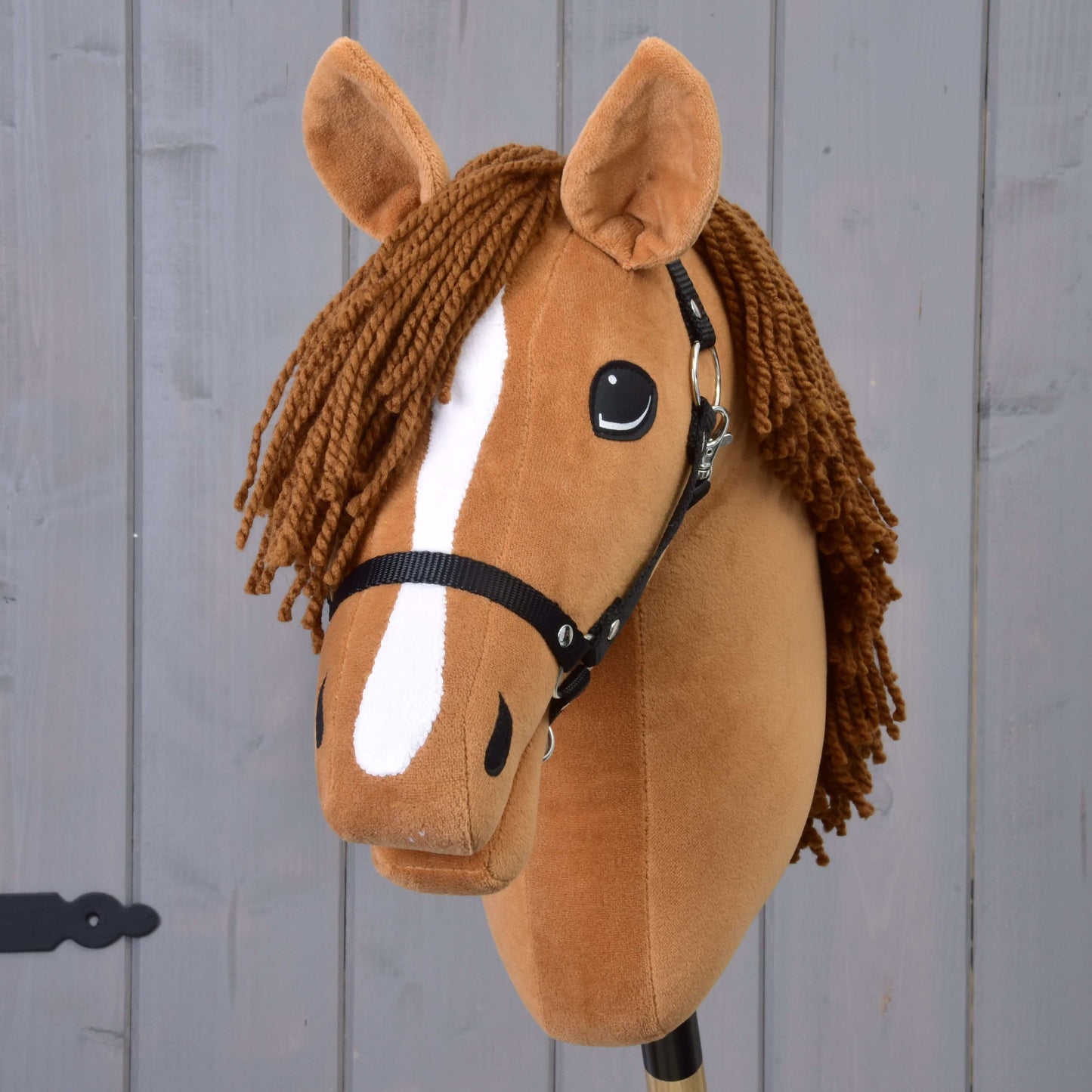 Hobby Horse Wendy s černou ohlávkou od Hobby Horse LarDen má hnědou plyšovou hlavu, spletenou hnědou hřívu z příze, bílý pruh na čenichu a černou ohlávku, inspirovanou ryzkou klisnou, a je zobrazená na šedém dřevěném pozadí.